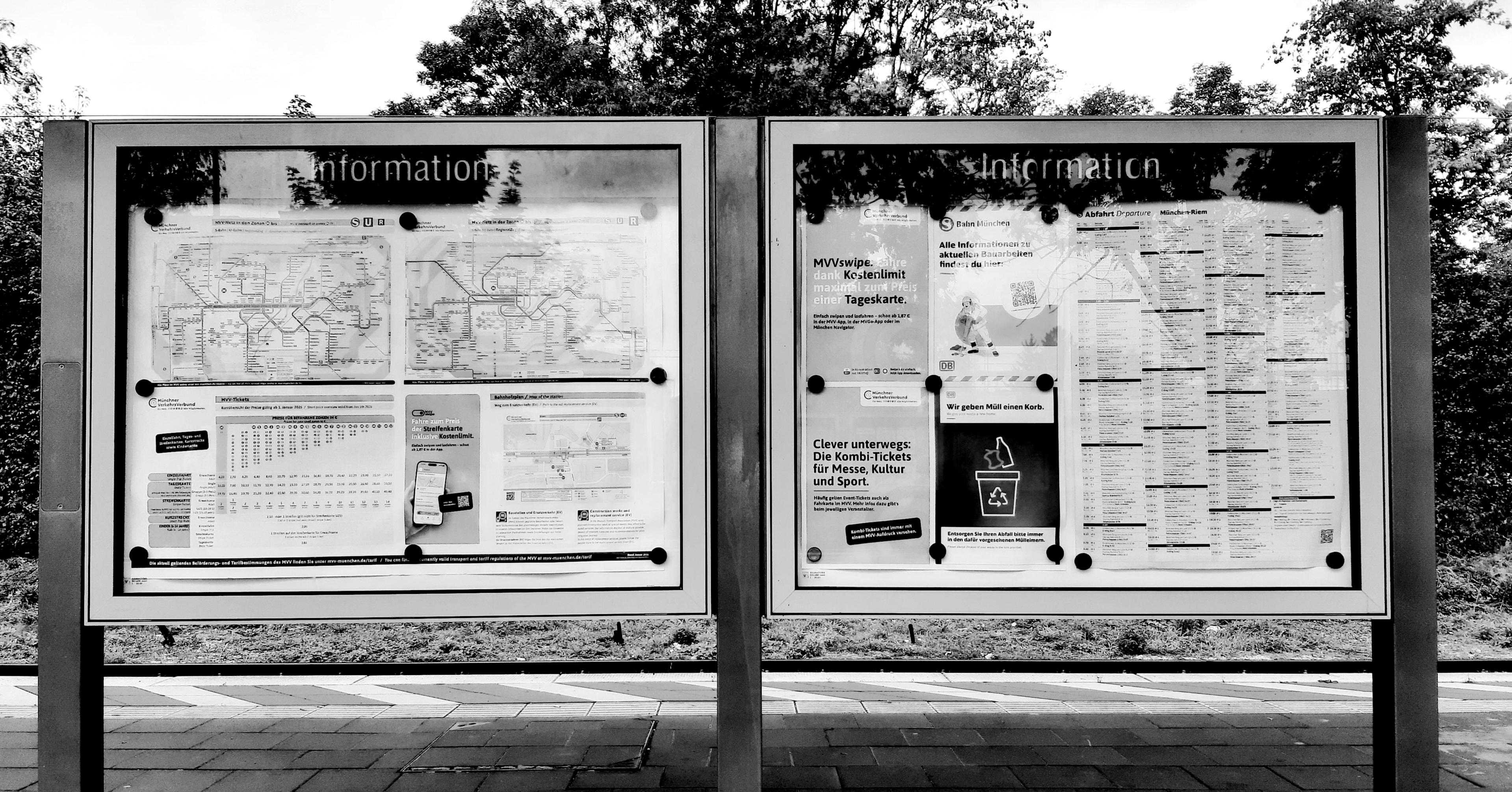 A double glass display, showing the regional railway network, a square meter of printed train schedule, a table with fares, and Posters withvuseful information. The picture is Black and white, it shows the marked edge of the platform, and a bit of the plants of the environment