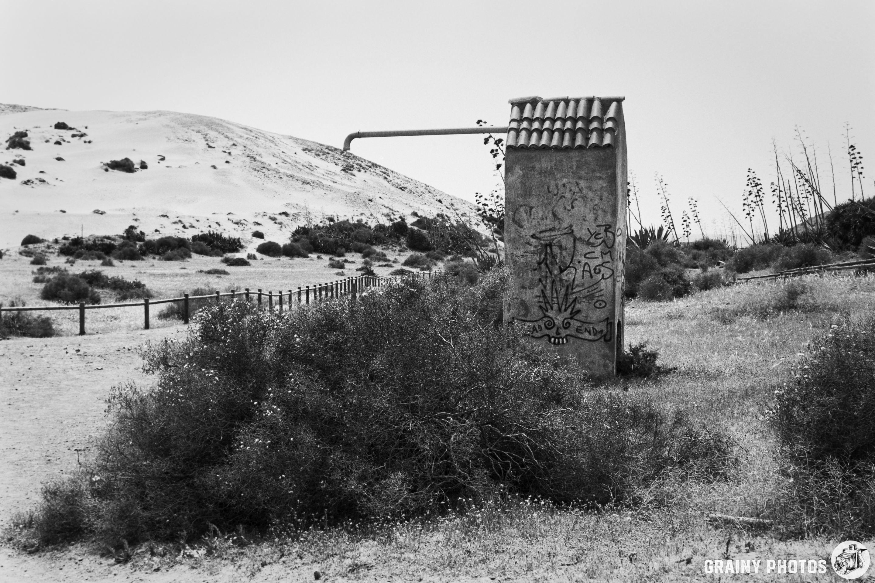 A weathered concrete structure with graffiti stands in a lush, grassy area beside a dusty trail, against a backdrop of rolling sand dunes and sparse vegetation. The scene is captured in black and white, evoking a sense of urban decay in a natural setting.