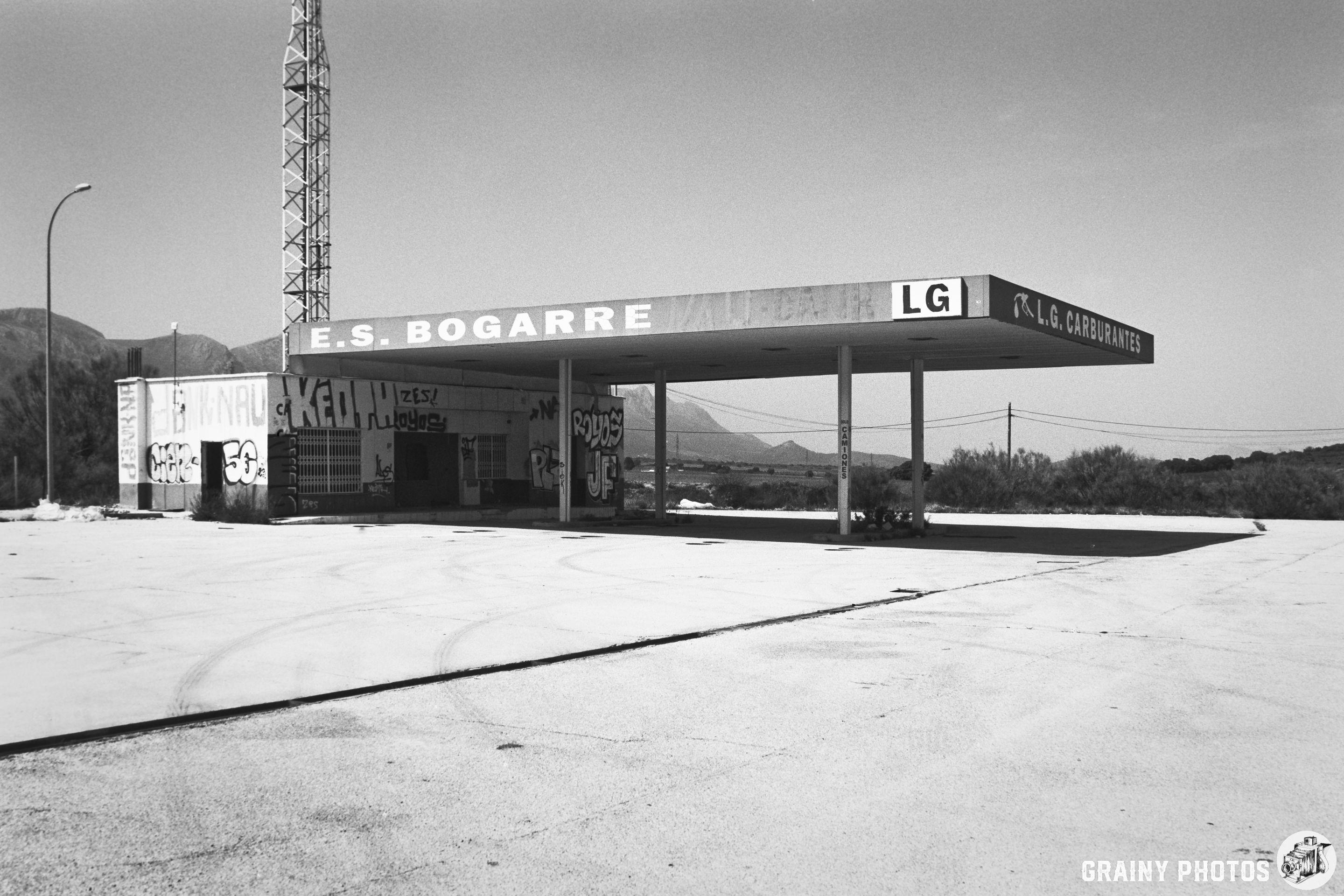 Abandoned petrol station with graffiti on the building and forecourt, surrounded by mountains and sparse landscape in a black and white setting.