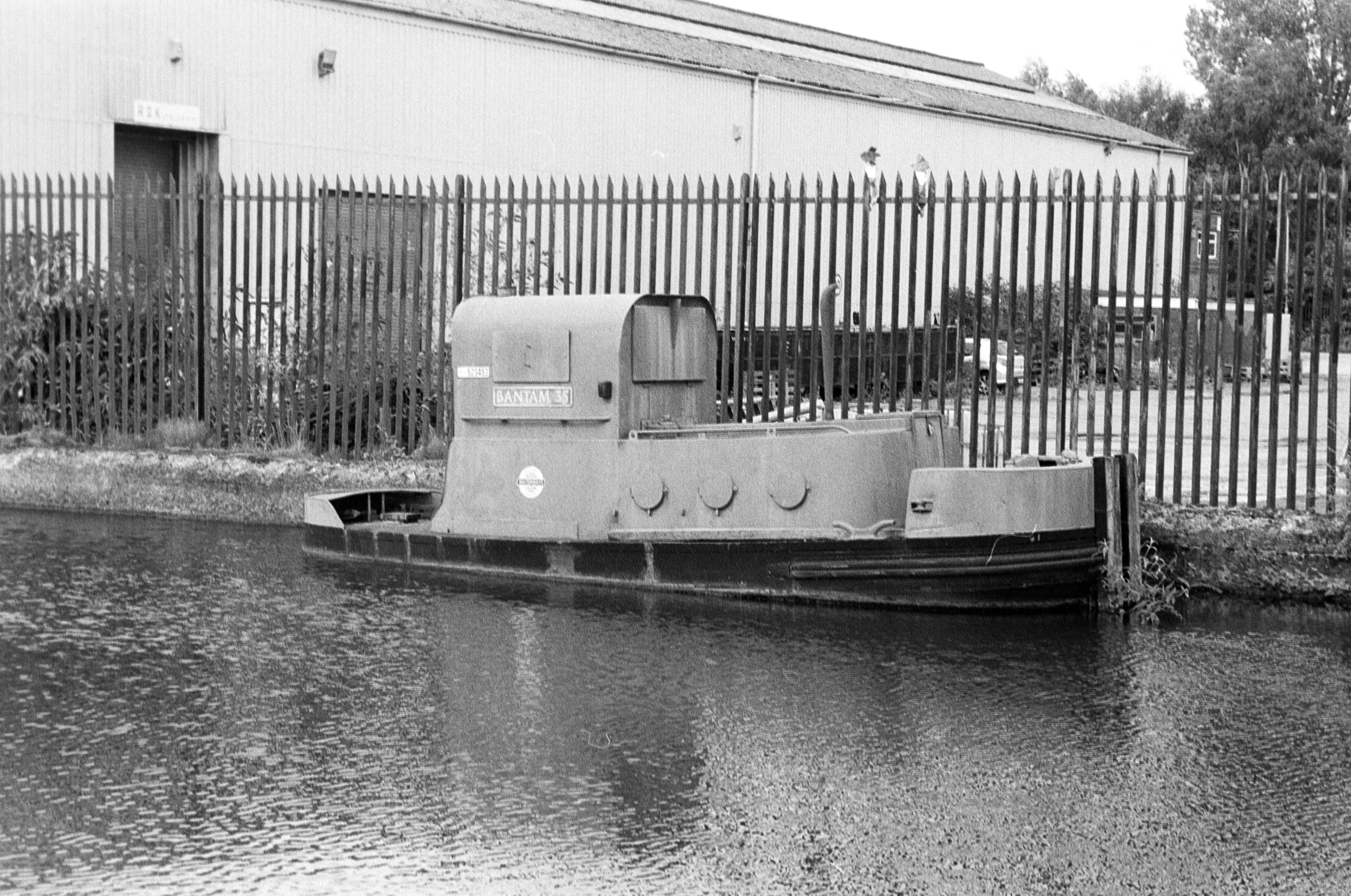 A black and white film photograph of Canal Tug Bantam on the Leeds & Liverpool Canal next to The Brick Community Stadium, Wigan.