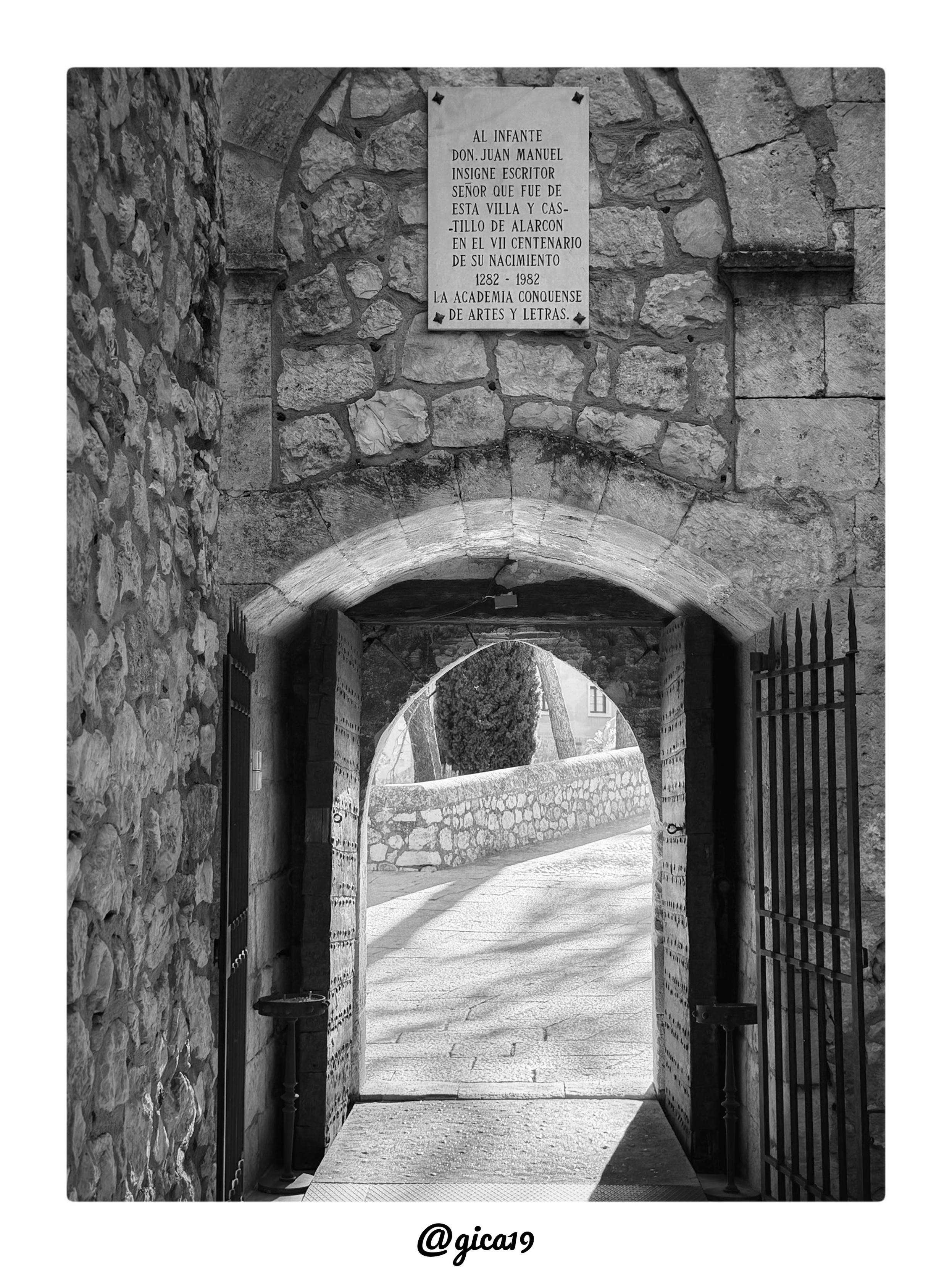 Esta es una fotografía en blanco y negro de la puerta de acceso al patio del Castillo de Alarcón, en Alarcón, provincia de Cuenca.

La imagen está tomada de frente, con simetría marcada, mostrando un vano de entrada abierto en un grueso muro de piedra. El muro está construido con sillarejo y mampostería irregular de distintos tamaños, característico de la construcción defensiva medieval. Sobre la puerta se dispone un arco de medio punto, formado por dovelas de piedra bien labradas que contrastan con la rusticidad del muro que lo rodea.
Encima del arco, incrustada en la pared, hay una placa conmemorativa de piedra con una inscripción dedicada al Infante Don Juan Manuel, escritor e insigne señor de la villa y castillo de Alarcón, colocada con motivo del séptimo centenario de su nacimiento (1282-1982) por la Academia Conquense de Artes y Letras.

La puerta en sí es de madera antigua, reforzada con remaches o clavos metálicos dispuestos en filas, y está flanqueada por dos hojas de verja de