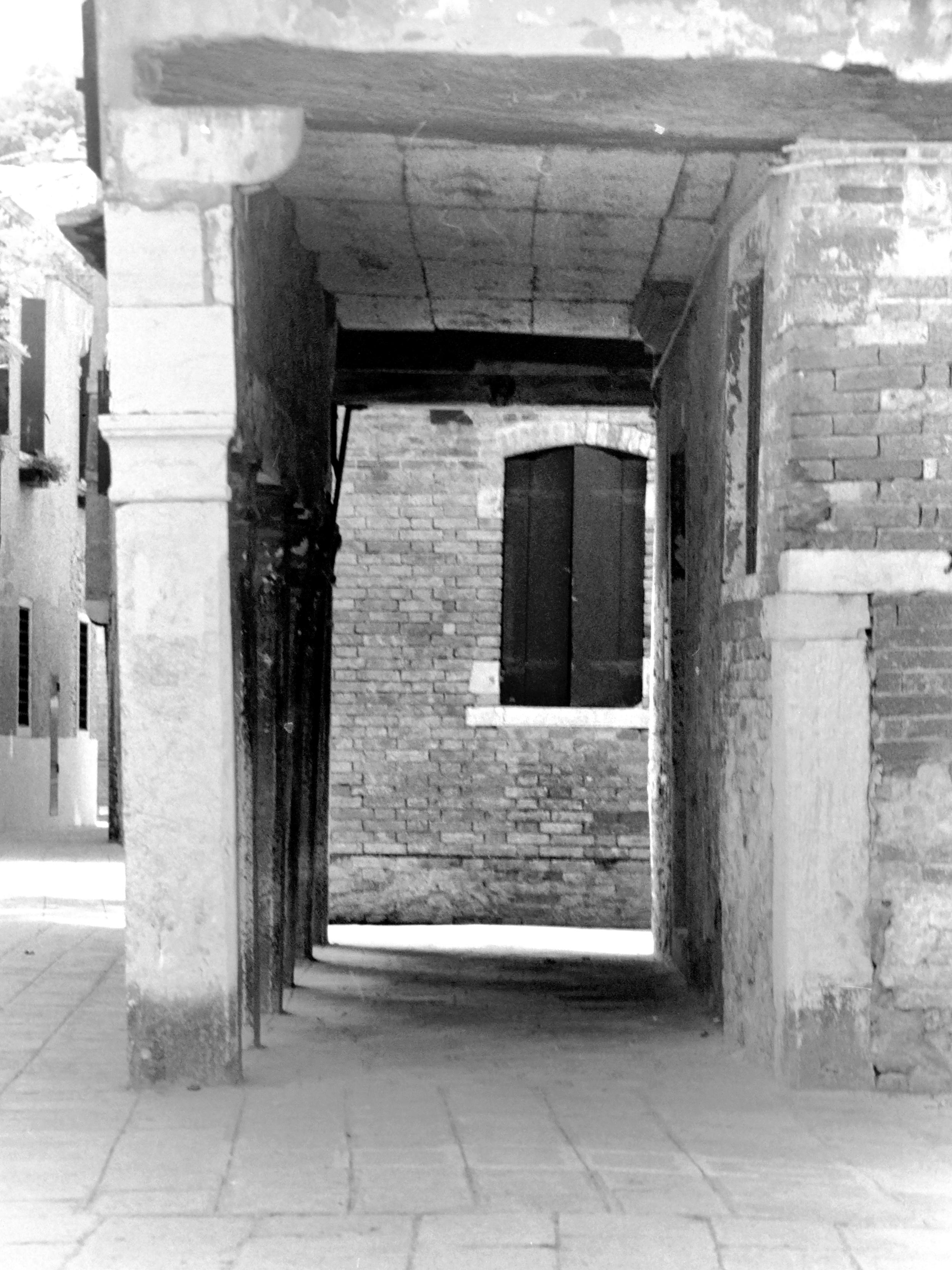 The "analog" photograph, in black and white and 3:4 portrait format, shows a window with closed shutters in a brick wall seen through a porch supported by a set of columns and a brick wall with light-colored stone reinforcements. On the left, a small section of street and facade with windows. The floor is tiled.