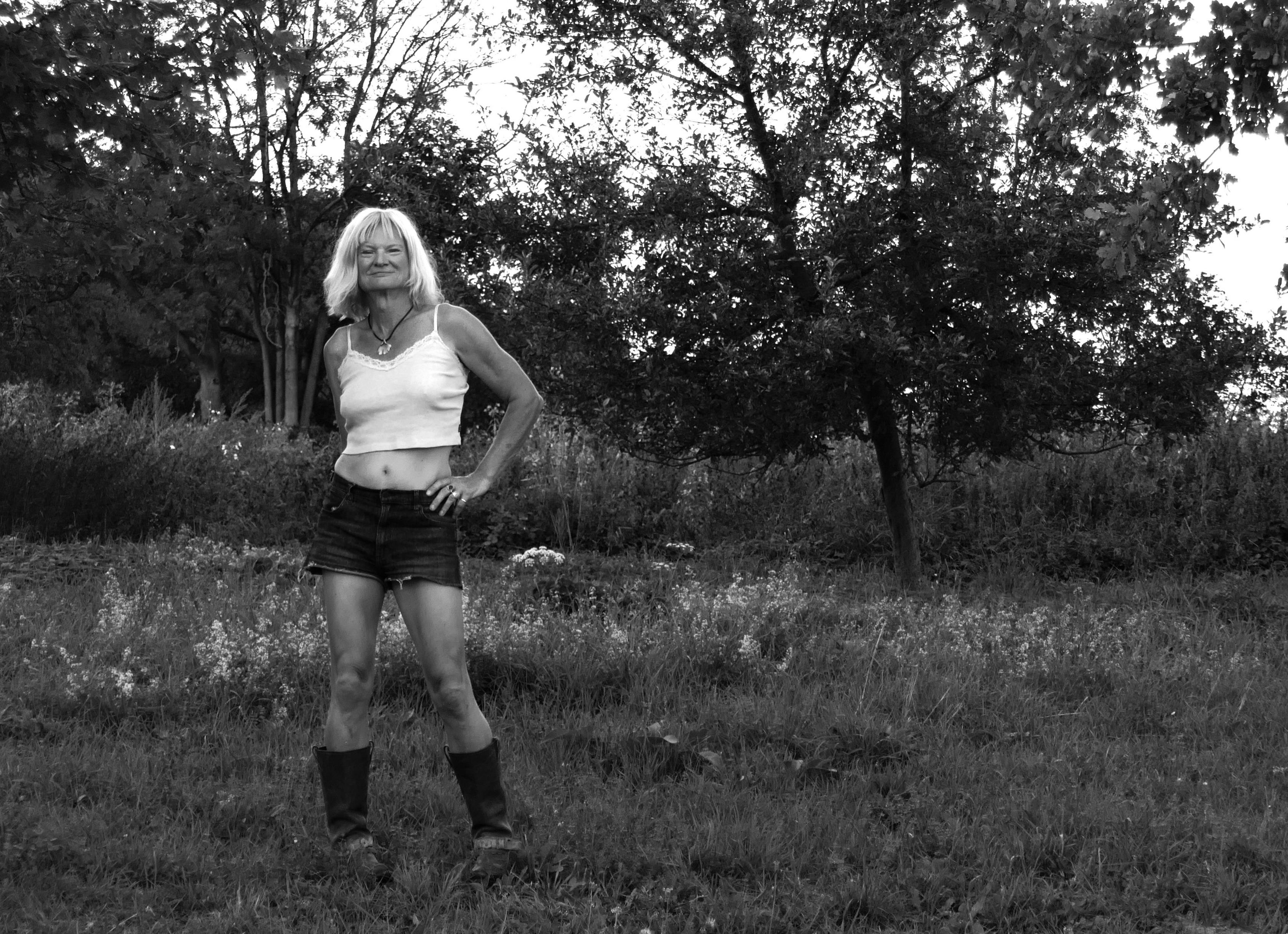 A woman on a meadow, she is rather small on the picture, standing left from the center. wearing shorts, a cropped top and harness boots. She faces the camera, her blond hair seems white. The grass and brush and small trees are dark, nearly black. A grey sky shines through the branches.