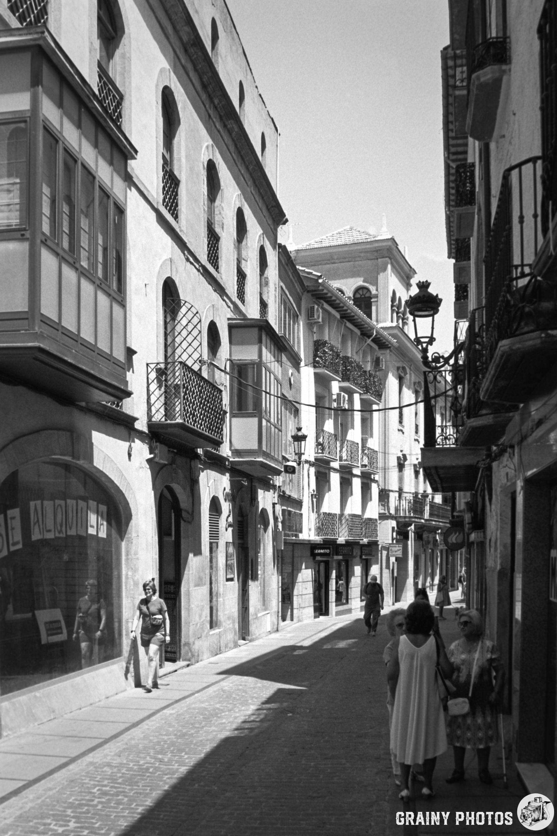 A narrow, sunlit street in Cazorla featuring traditional architecture, balconies, and pedestrians. Black-and-white photo captures a sense of timeless charm with people walking and exploring the vibrant urban atmosphere.