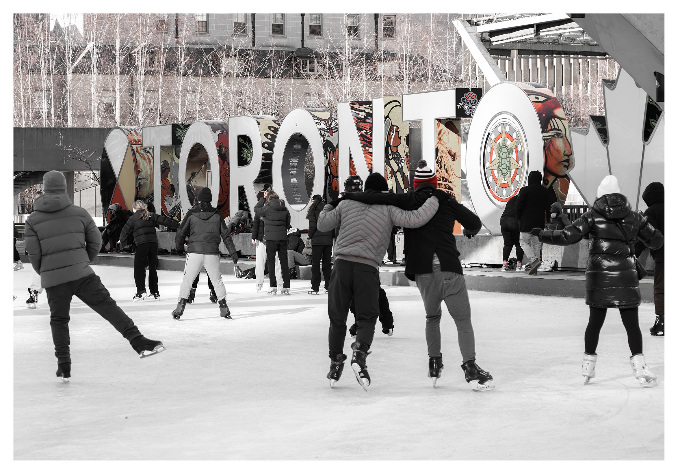 A photo of people skating, alone or with friends, in front of the Toronto sign at the ice rink in front of Toronto's city hall. The image has been desaturated with the exception of the Indigenous art on the large letters making up the sign and the toque of one of the skaters. In front of those colours, the skaters, experienced and inexperienced, enjoy the feeling of gliding on the ice. In the background, thin, white winter trees with naked branches and a city building.