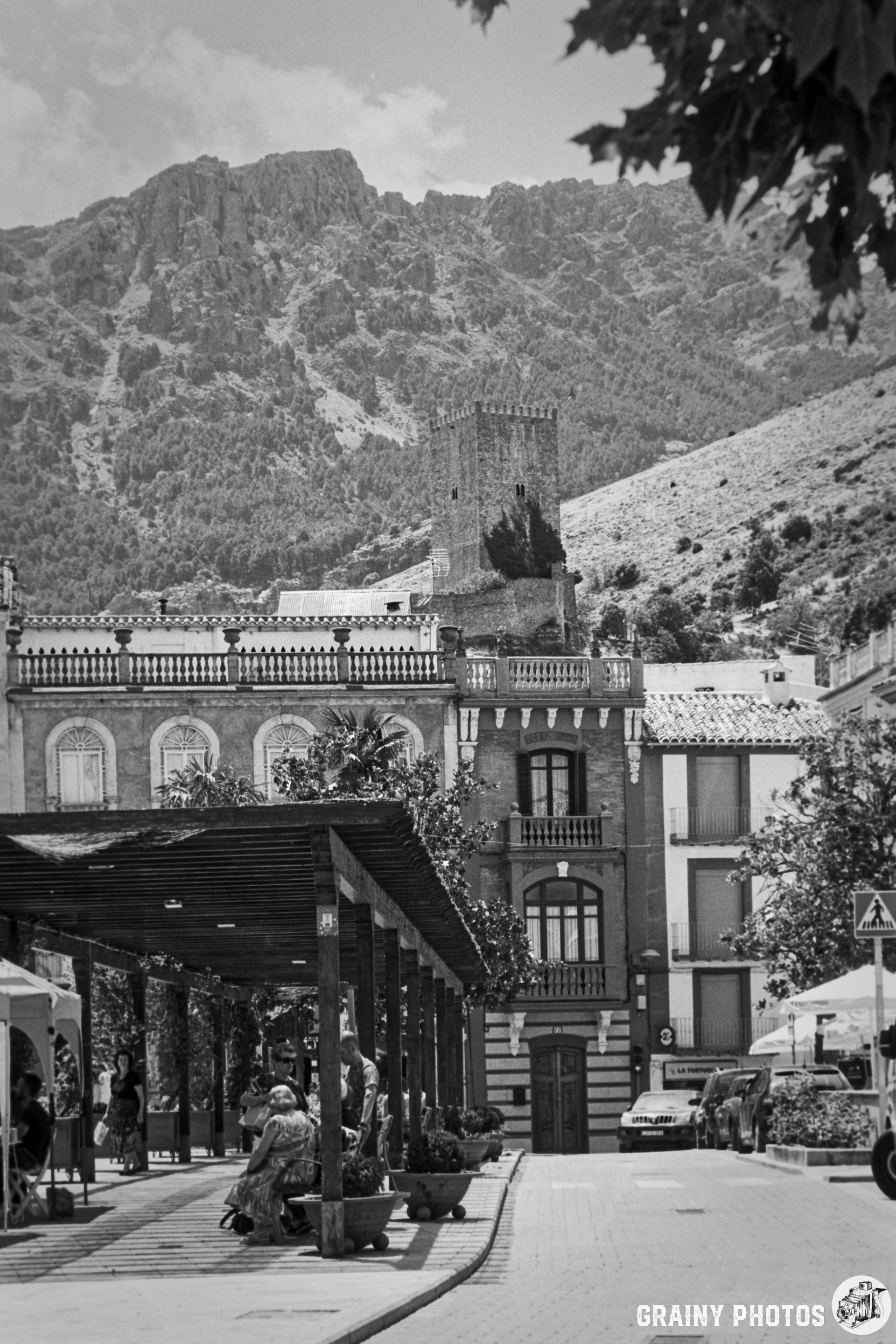 A black and white view of one of the three squares in Cazorla, lined with trees and benches, featuring historic buildings and a mountainous backdrop, showcasing a blend of culture and nature.
