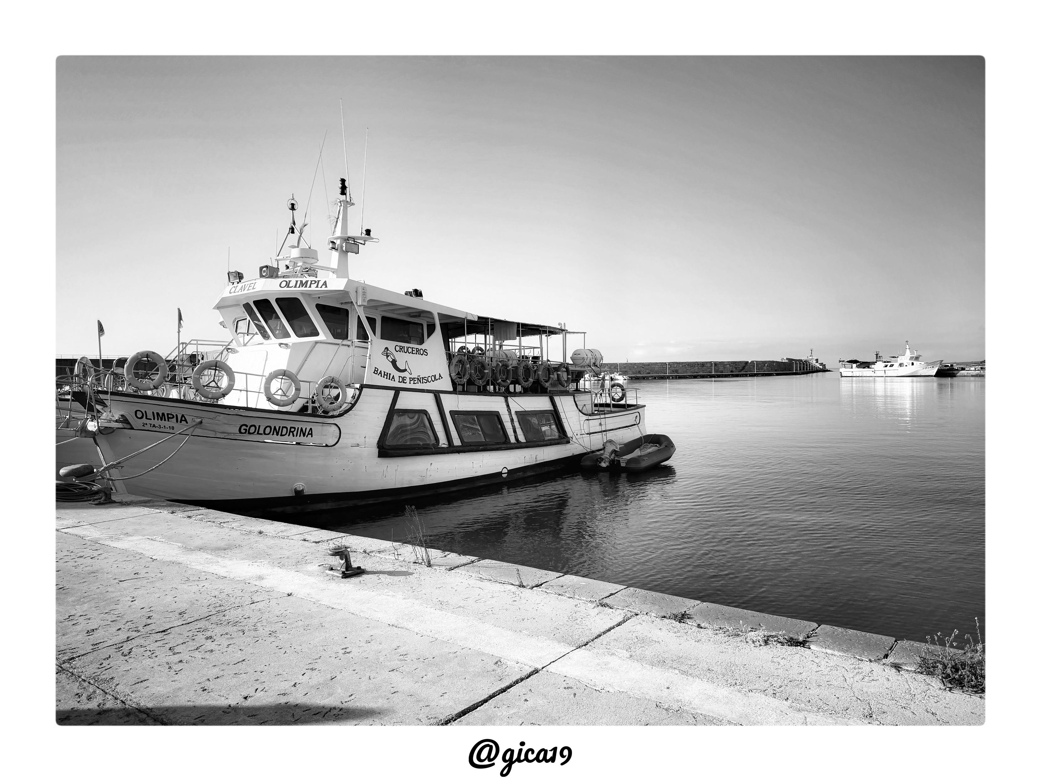 La fotografía, en blanco y negro, muestra una escena portuaria tranquila en el Puerto de Peñíscola durante las primeras horas de la mañana.

El encuadre está dominado en primer plano por un muelle de hormigón vacío, con las juntas de las losas marcando líneas diagonales que conducen la mirada hacia el agua. La superficie del muelle presenta manchas y una pequeña grieta con hierba silvestre asomando, además de una argolla metálica de amarre anclada en el borde. El agua del puerto aparece completamente en calma, casi como un espejo, reflejando tenuemente las embarcaciones y el cielo despejado.

El elemento central de la imagen es un barco de pasaje de tamaño medio, amarrado de proa hacia el muelle. En su casco se leen los nombres “Olimpia” y “Golondrina”, junto con una matrícula. En la parte superior de la cabina se distingue el rótulo “Cruceros Bahía de Peñíscola” con un pequeño logo de un pez o delfín, lo que indica que se trata de una embarcación dedicada a excursiones turísticas por 