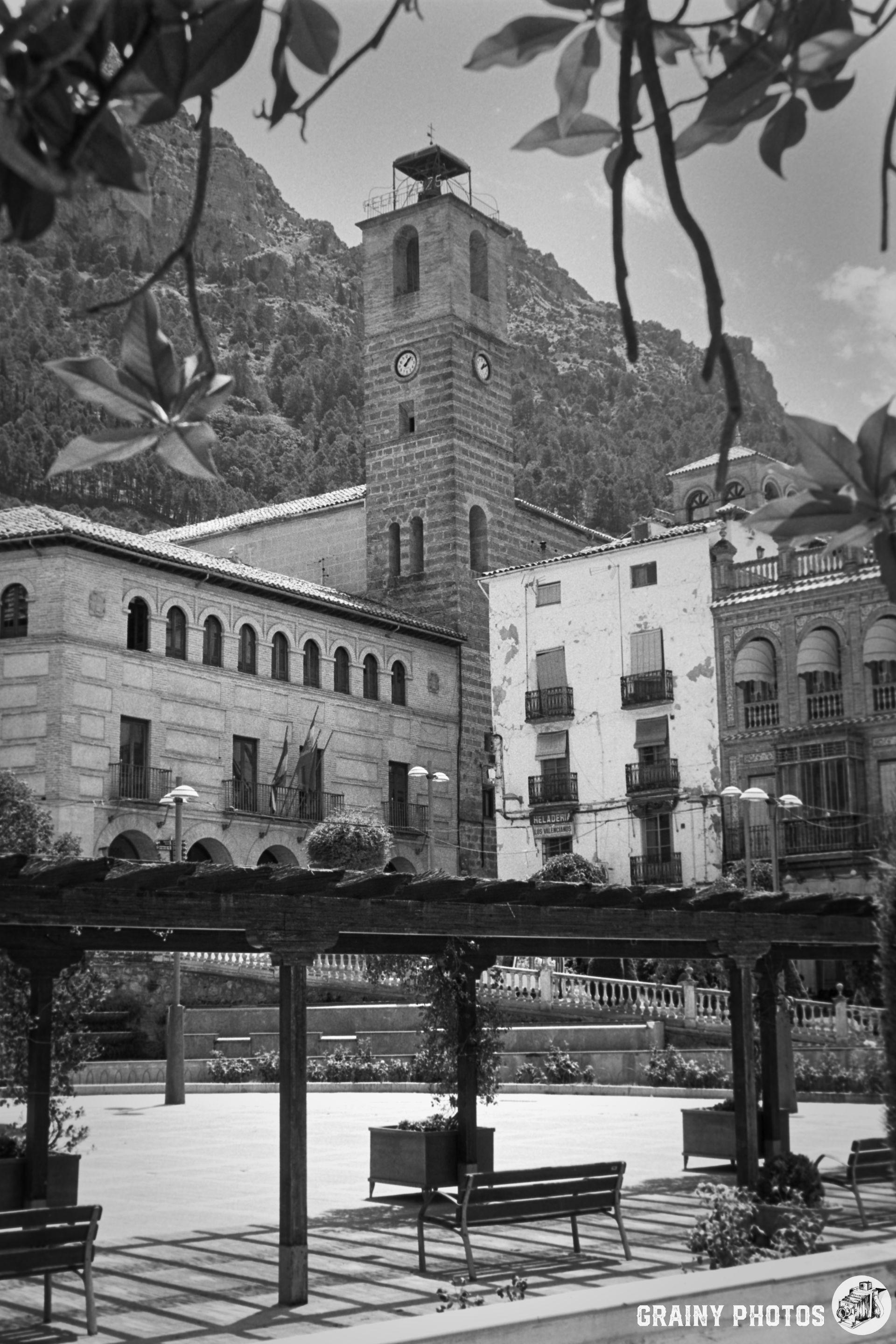 A black and white photo of a historic town square featuring a clock tower, surrounded by traditional architecture and a mountainous backdrop. The foreground includes wooden benches and trellises with greenery, creating a serene atmosphere.
