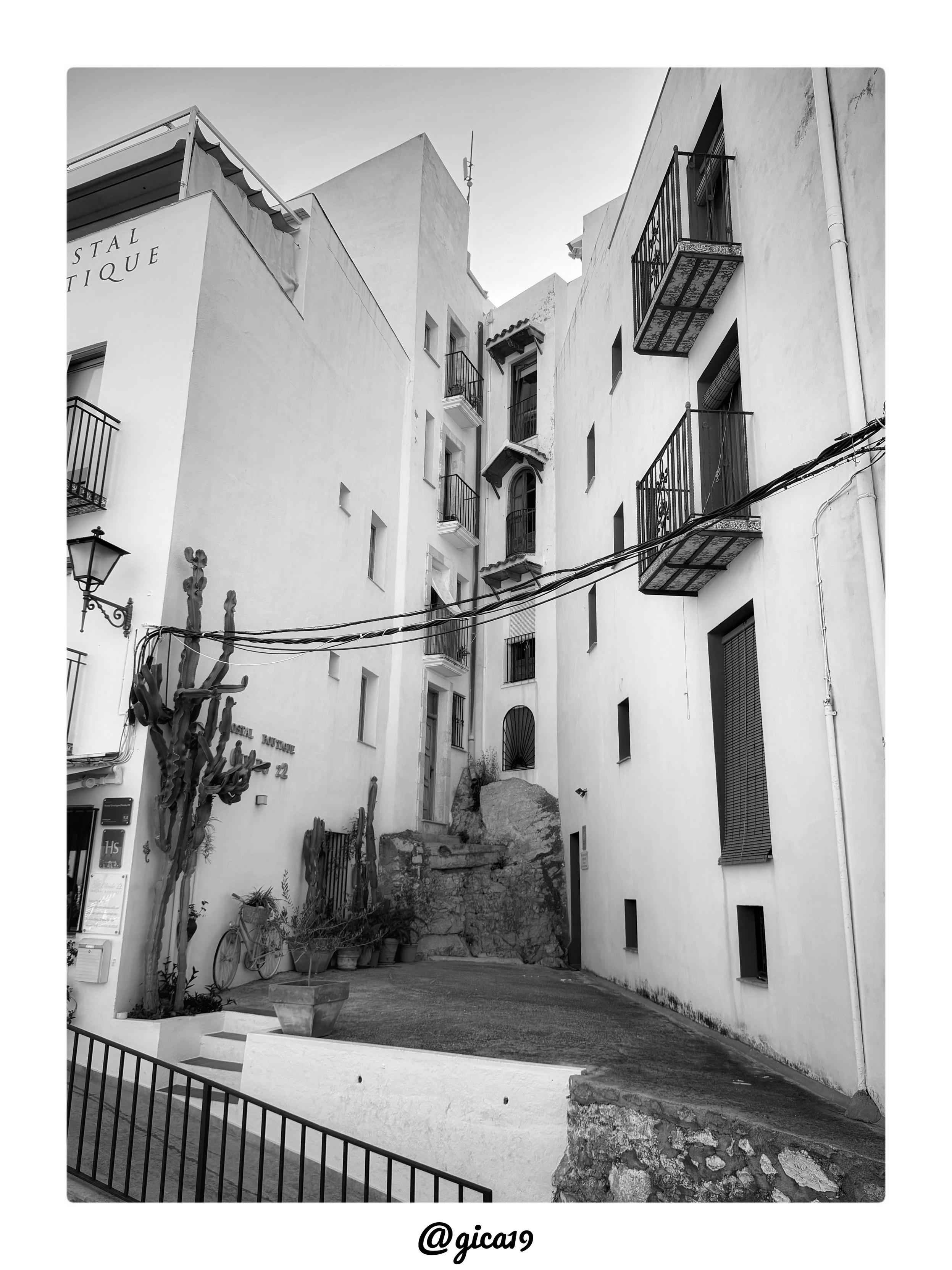 La imagen es una fotografía en blanco y negro, tomada en una de esas callejuelas empedradas y estrechas que uno se encuentra sin buscarlas en el casco antiguo de Peñíscola. Es de mañana, se nota enseguida: la luz llega suave, casi tímida, filtrándose entre los edificios como si aún no se hubiera despertado del todo.

La toma está en vertical, mirando hacia arriba, por un callejón tan angosto que casi se puede tocar una fachada con cada mano. A la izquierda se levanta un edificio de varias plantas con el rótulo de un hostal, cuyo nombre solo se adivina a medias en la parte superior —el tiempo y la luz se han encargado de difuminarlo un poco—. Al fondo, y también a la derecha, aparecen otras construcciones blancas, de alturas distintas, y es que ahí está la gracia: se genera esa sensación de laberinto vertical tan propia de las calles peñiscolanas, donde nunca sabes muy bien hacia dónde te va a llevar el siguiente giro.

Lo que más llama la atención son los balcones, con sus barandillas 