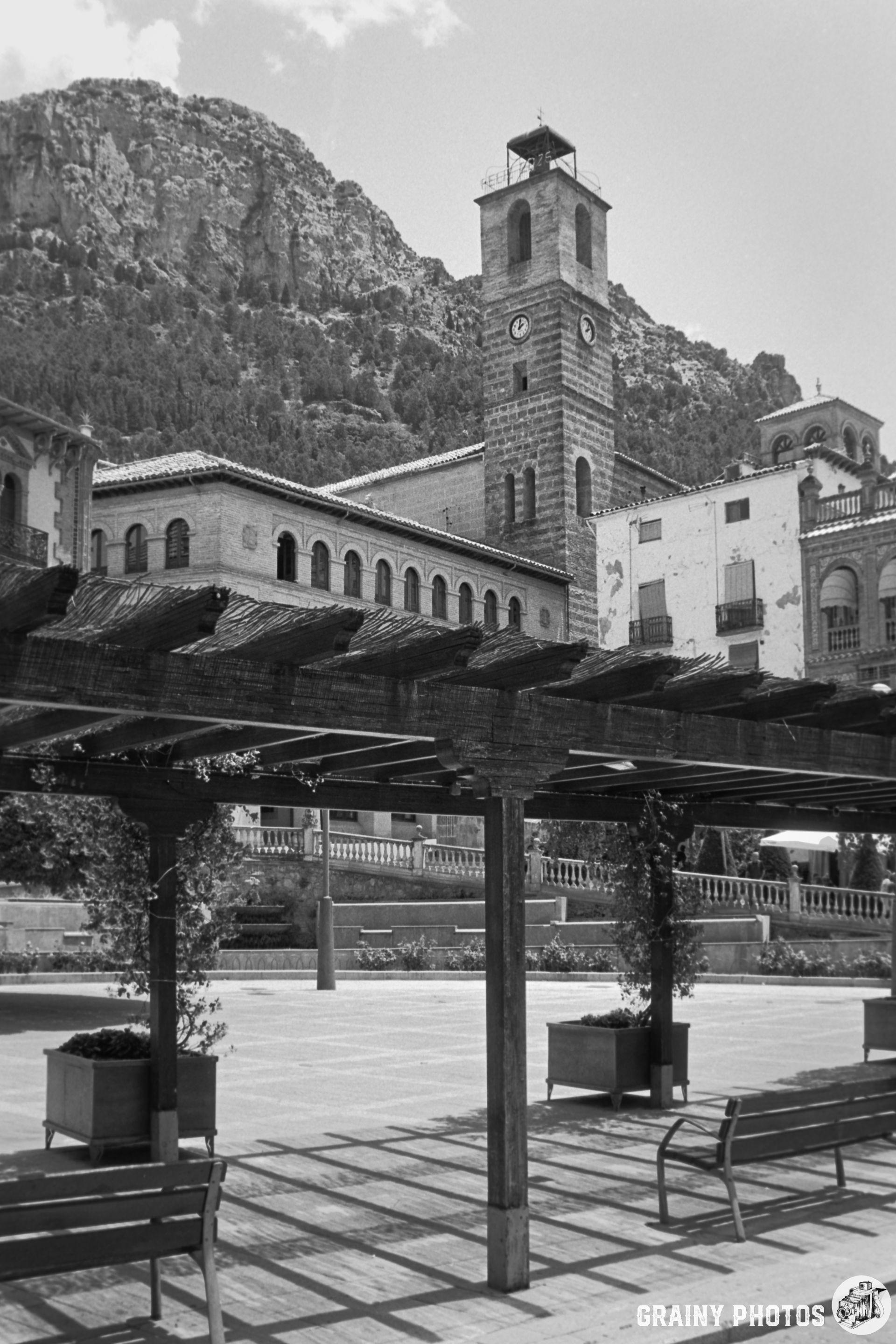 A scenic black and white view of Plaza de la Corredera in Cazorla featuring a clock tower, historic buildings, and a pergola with benches, framed by a mountainous backdrop.