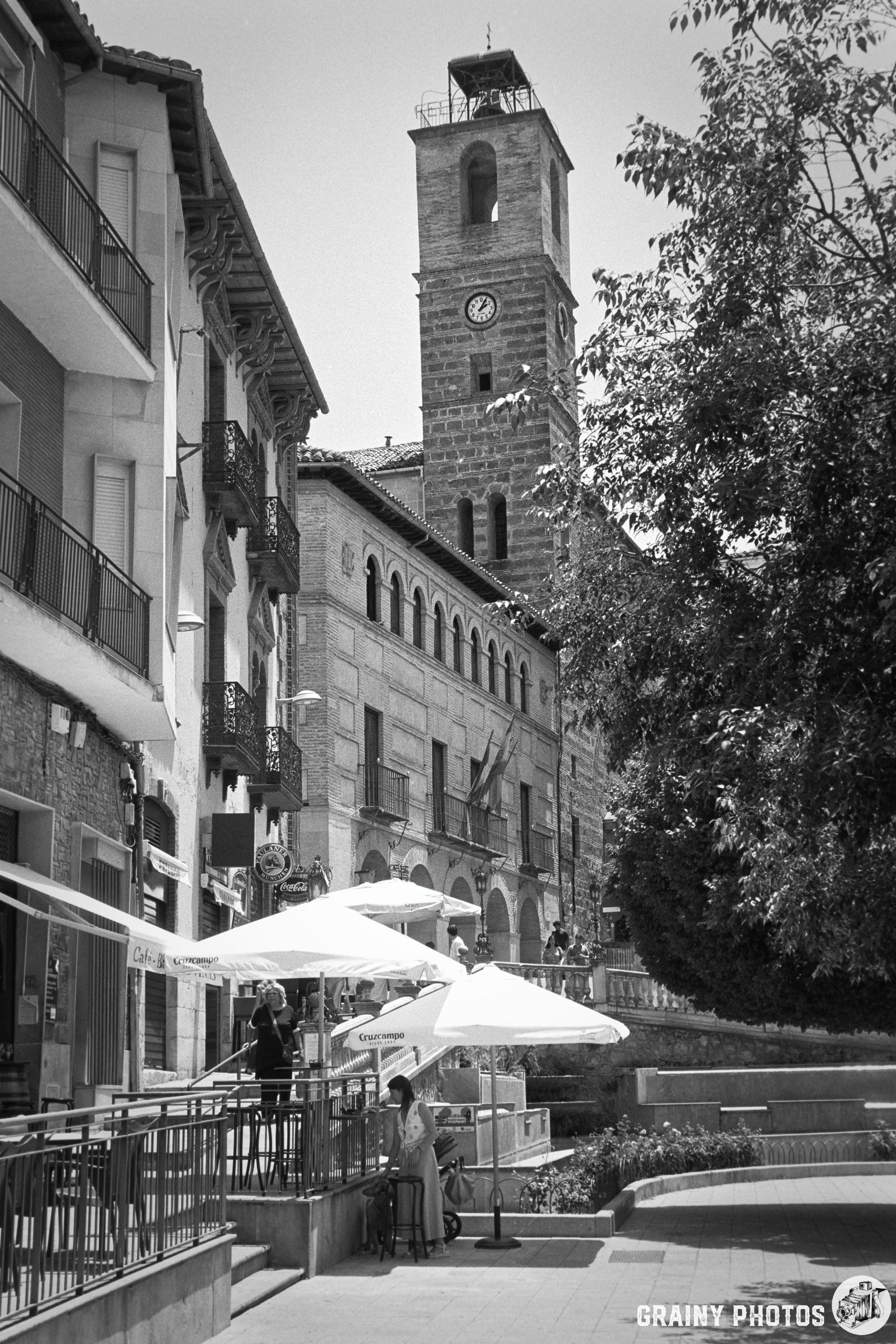 A black-and-white image of a quaint street scene featuring a clock tower, outdoor cafes with umbrellas, and people enjoying the atmosphere. The architecture showcases historical buildings alongside a tree-lined pathway.