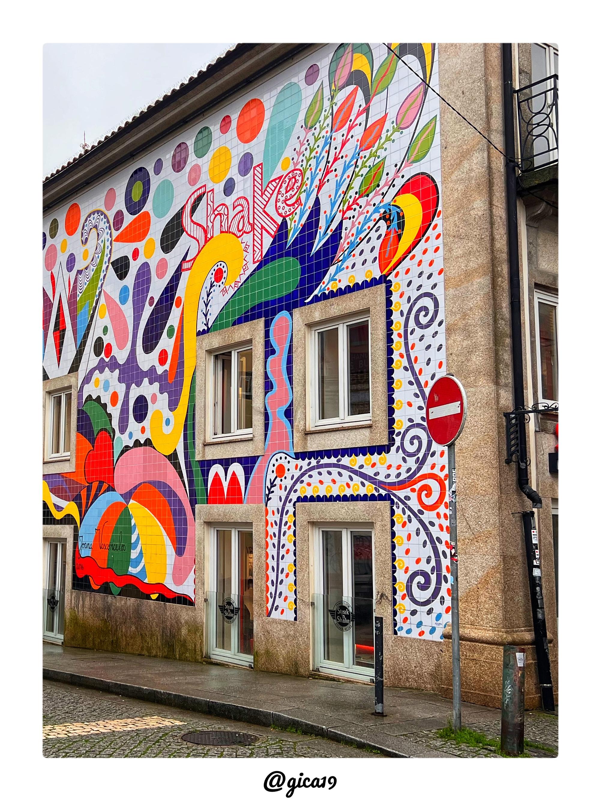 Esta imagen muestra un espectacular mural de azulejos que cubre la fachada lateral de un antiguo edificio en la Praça Guilherme Gomes Fernandes, en el barrio de Cedofeita, en Oporto, muy cerca de la Torre de los Clérigos. Se trata de la obra de la artista portuguesa Joana Vasconcelos, creada en 2016 e inaugurada a comienzos de 2017 para la fachada del restaurante Steak ’n Shake.

El mural está formado por unos 8.000 azulejos pintados a mano y se extiende a lo largo de aproximadamente 20 metros, convirtiéndose en una de las obras de arte público más grandes creadas por la artista. La fachada del edificio, de estilo tradicional con muros de granito visibles en los laterales, está completamente revestida por un diseño explosivo de formas orgánicas y geométricas: círculos de gran tamaño en tonos morado, rosa, turquesa, verde y naranja se agrupan en la parte superior izquierda, mientras que hacia el centro y la derecha se despliegan motivos que evocan hojas, plumas y formas florales estiliz