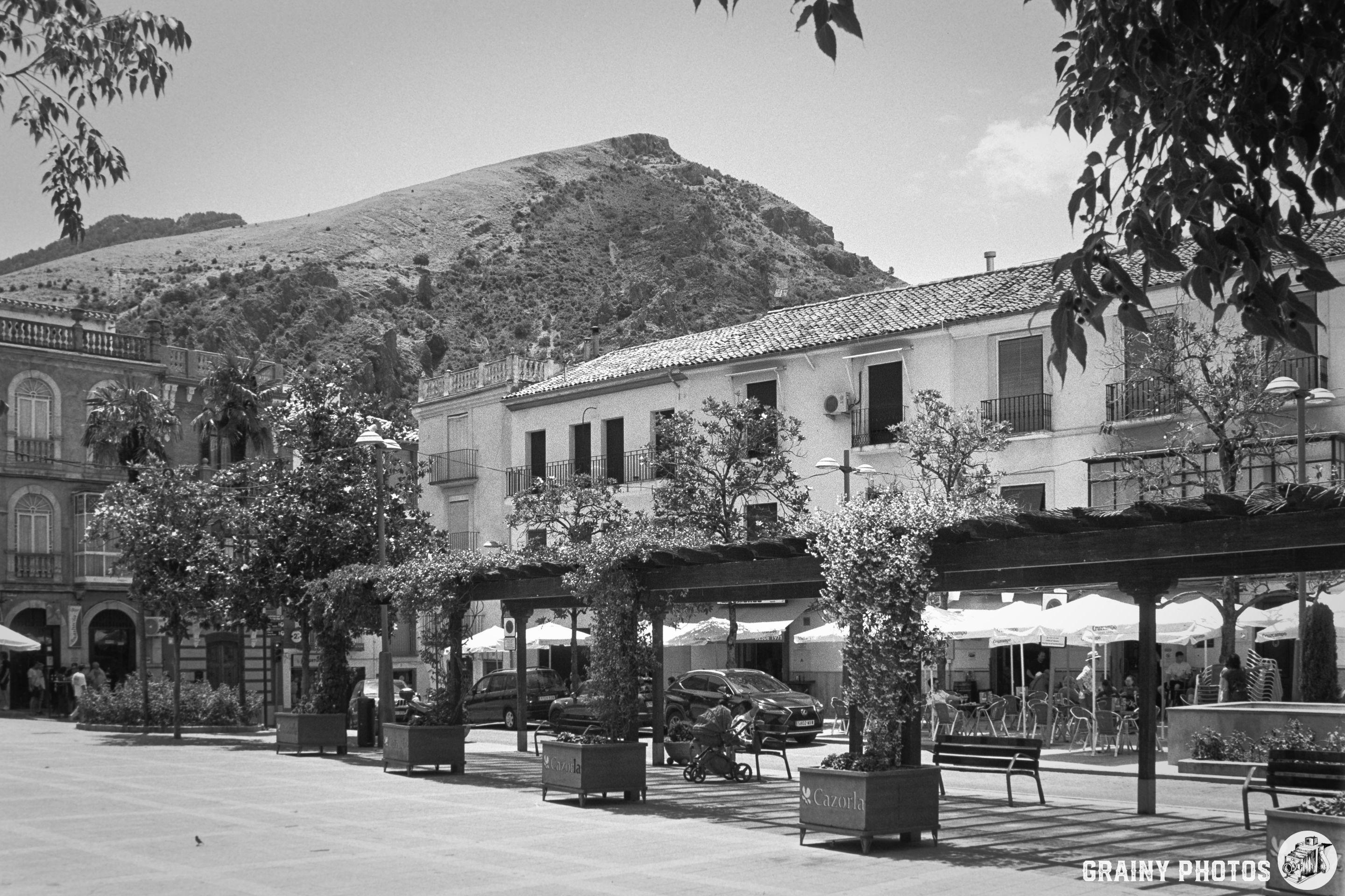A charming plaza with outdoor seating and trees, framed by buildings and a hillside in the background. The scene is depicted in black and white, adding a timeless quality to the lively atmosphere.