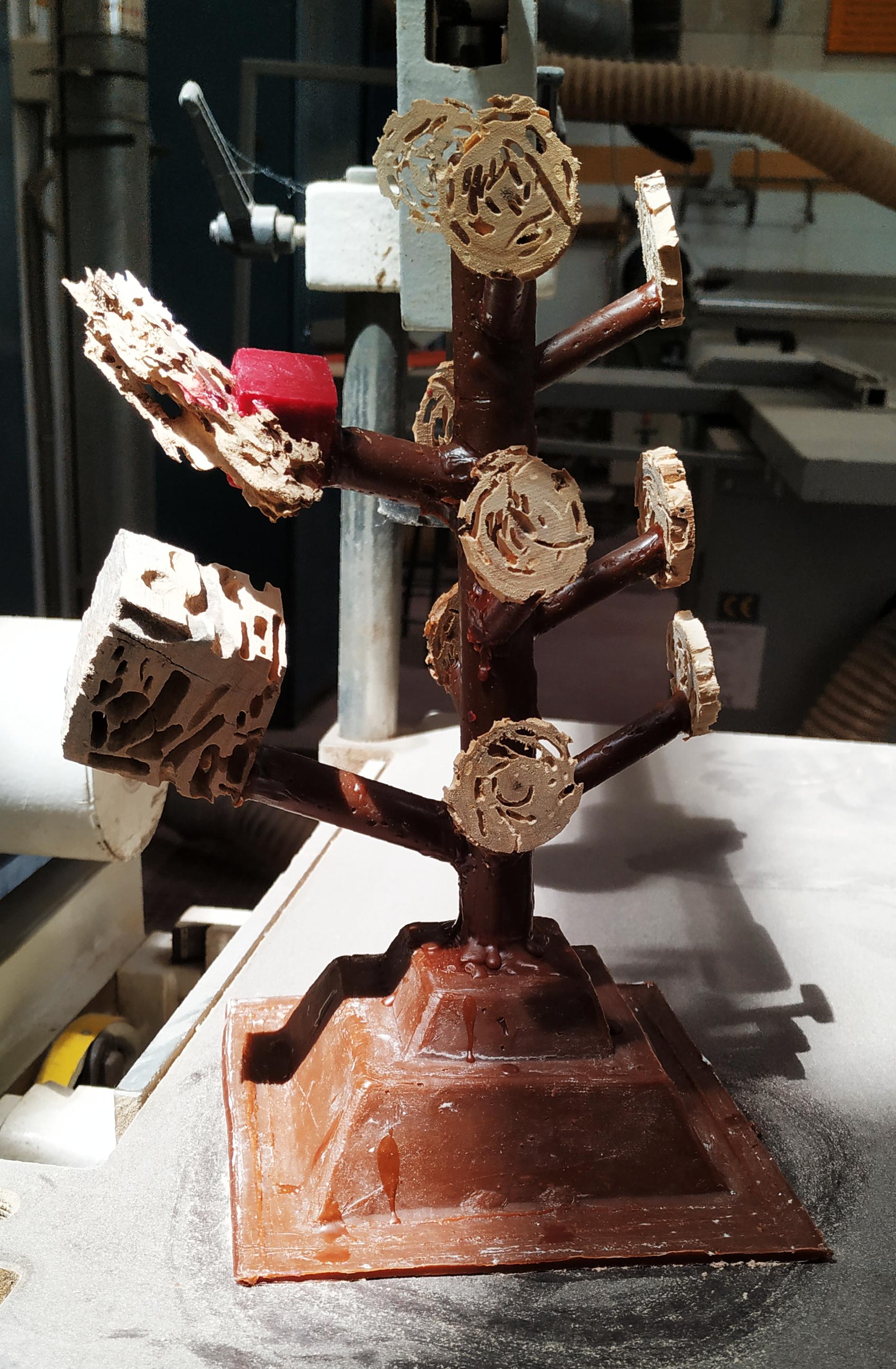 Circular slices of the handle, showing termite tunnels, and the cube from the previous post. They are all mounted on a red wax "tree". Everything will be covered in several baths of ceramics, making one fire resistant, hollow mold. The mold will be put on a kiln until the wax and wood turn to ash, leaving internal space for the liquid metal to be poured and conducted to the pieces.

The tree is upside down, it rests on its widest end, a pyramid shape where the metal will enter. From the tip of the pyramid, a central cylinder goes straight up, with smaller cylinders branching out of it, each one connecting to a wooden piece.