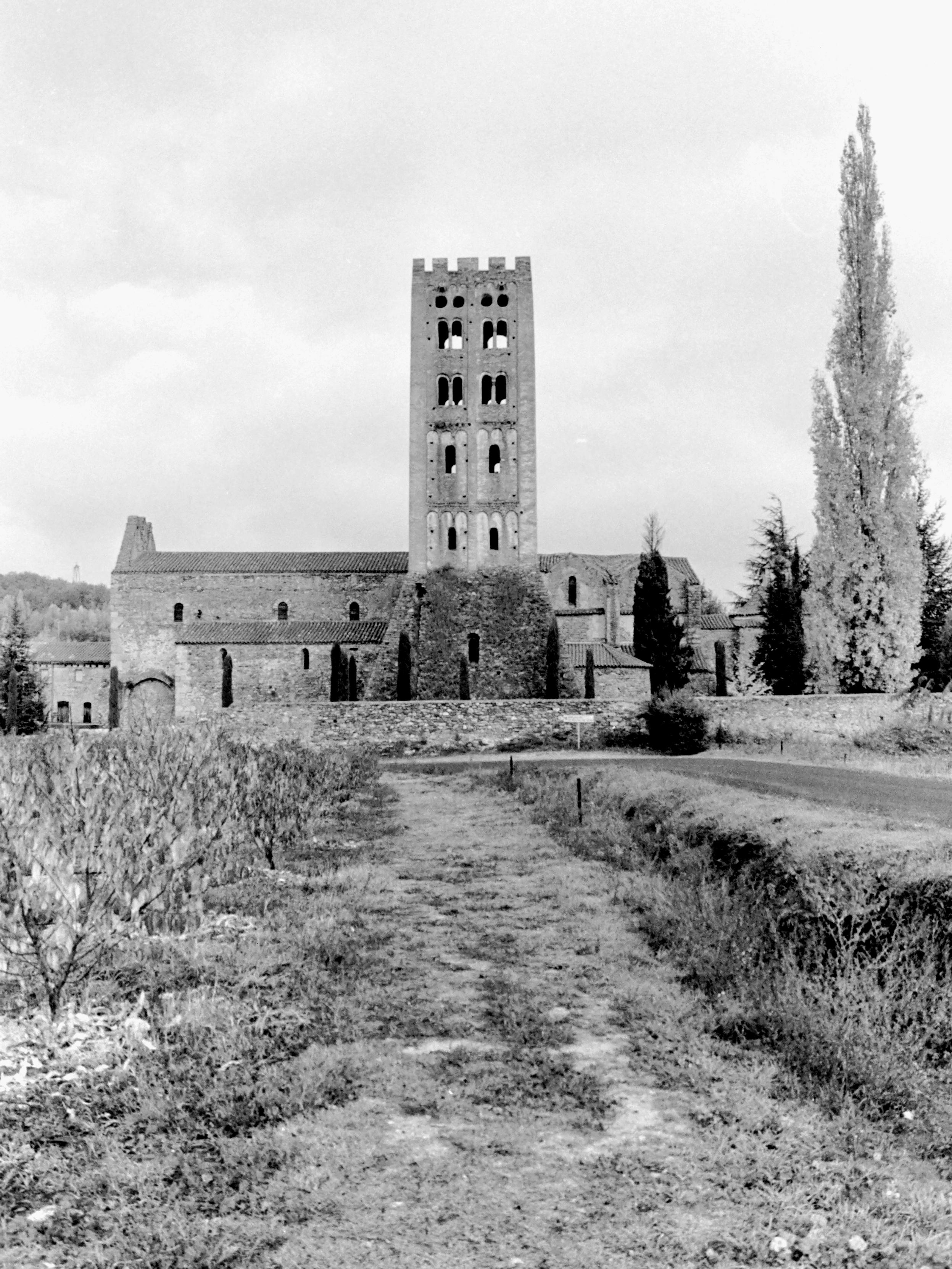The black and white analog photograph  shows a general view of the Benedictine Abbey of Sant Miquel de Cuixà, surrounded by trees, as seen from the adjacent countryside. The bell tower, a beautiful example of a Lombard-style Romanesque tower, stands out in the image. It consists of four floors that rise on a large late sloping base. The openings are distributed along the four levels: the first and second floors have two simple semi-circular arch windows on each side; on the third and fourth, also on each facade, there is a pair of twin semi-circular arch windows, separated by a column with a smooth capital and, finally, above the windows of the last floor there are small portholes.