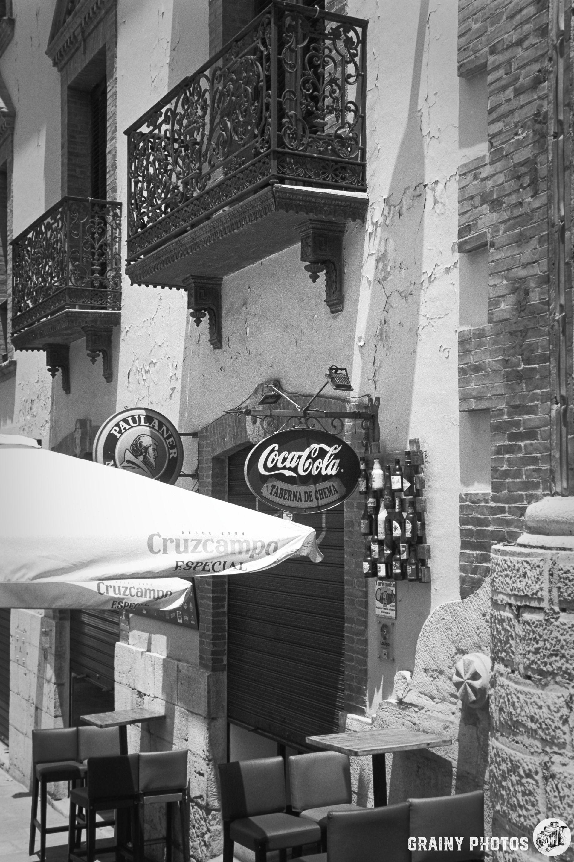 A historic building facade featuring two wrought iron balconies and a vintage Coca-Cola sign. In the foreground, an outdoor seating area with empty chairs and vibrant umbrellas adds to the inviting ambiance of this quaint locale.