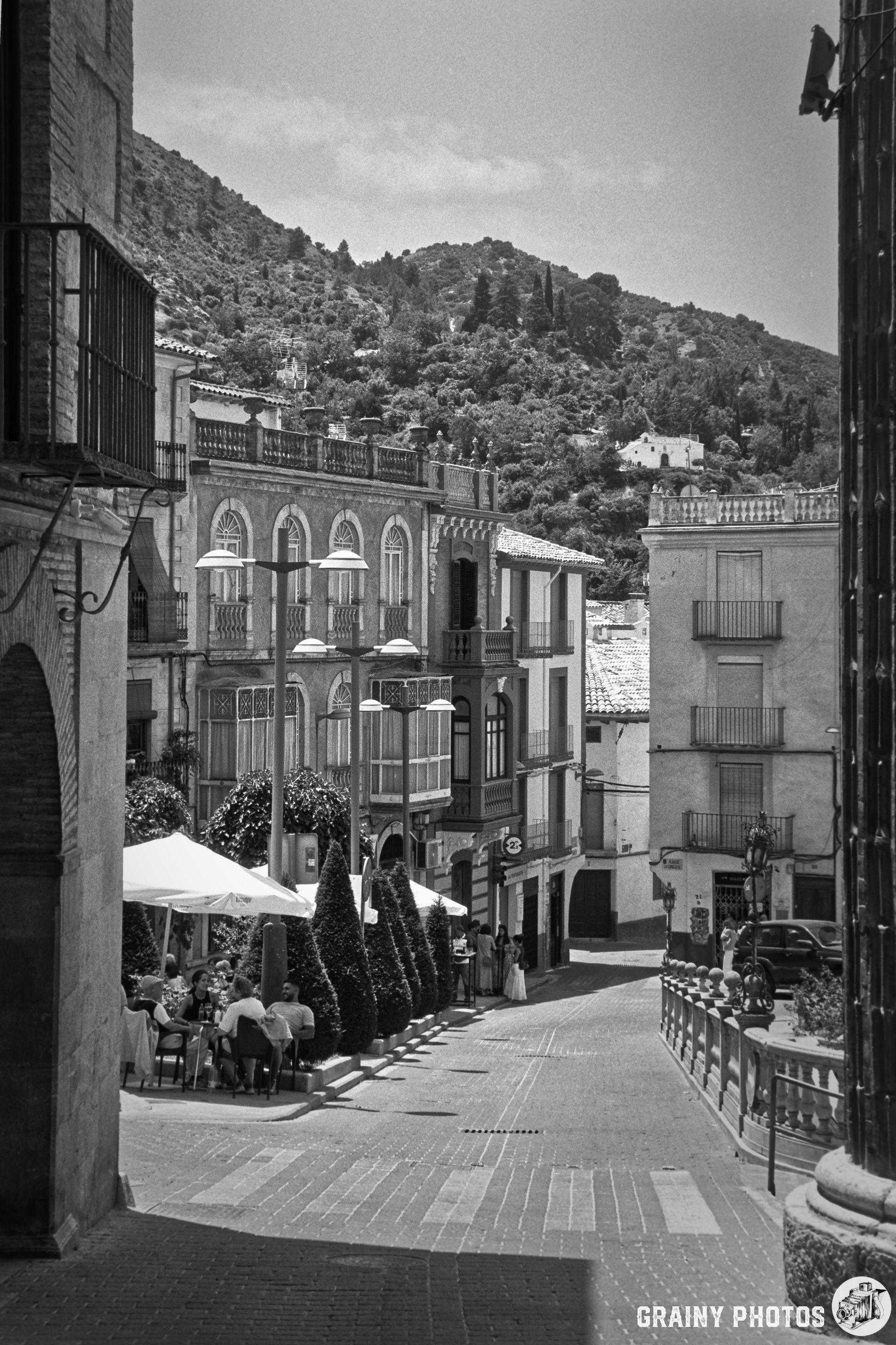A picturesque, narrow street in a Cazorla features traditional buildings, outdoor seating at cafés, and lush greenery on a sunny day, all set against a backdrop of hills. The scene conveys a charming, relaxed atmosphere.