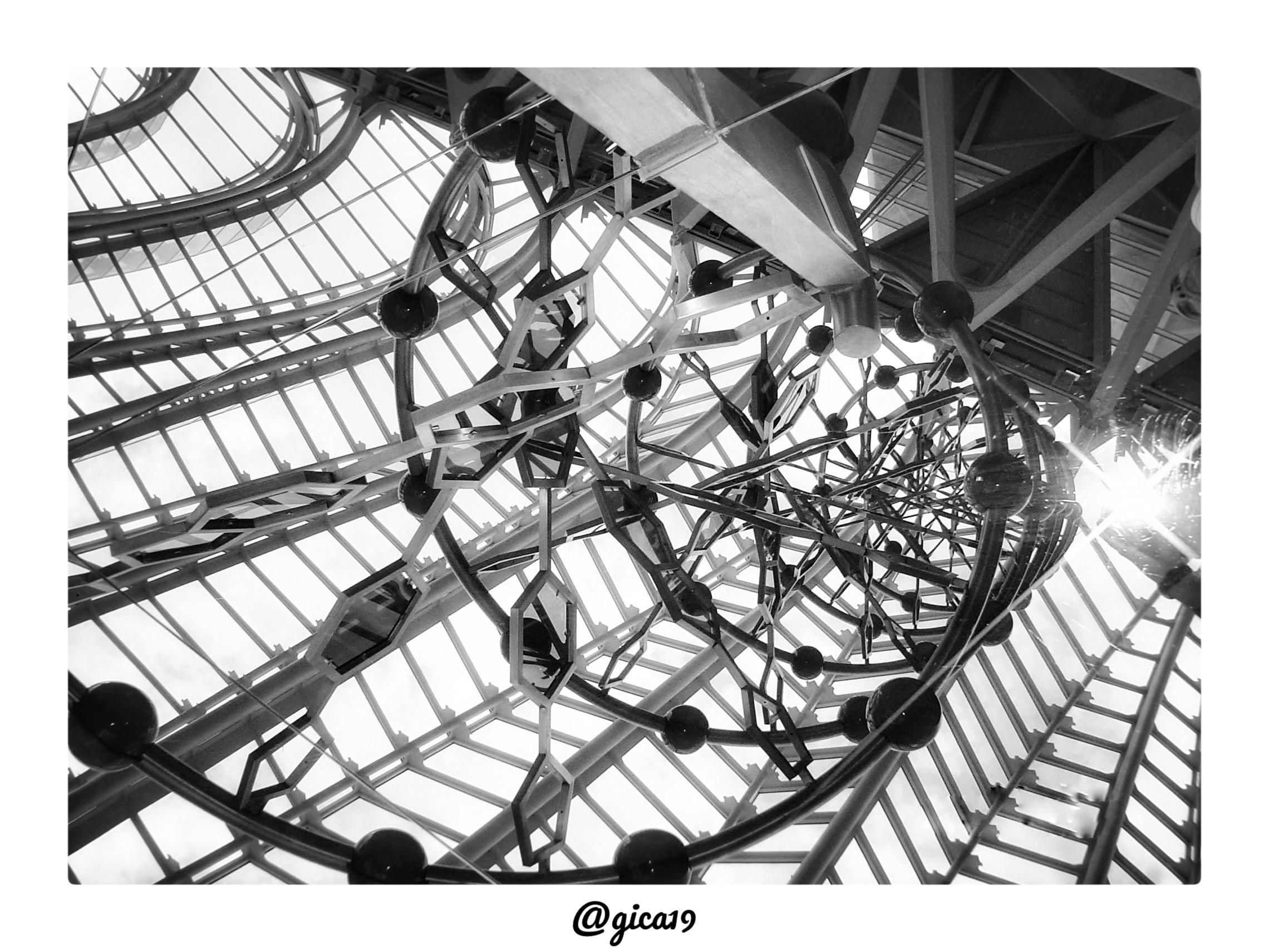 La fotografía en blanco y negro capta, desde un ángulo contrapicado, el interior del Museo de las Ciencias Príncipe Felipe de Valencia, obra del arquitecto Santiago Calatrava, integrada en la Ciutat de les Arts i les Ciències. La composición se centra en la mirada hacia arriba, hacia la gigantesca cubierta acristalada que corona el edificio, formada por una retícula de vigas metálicas triangulares y rómbicas que se extienden en diagonal por todo el encuadre, dejando pasar la luz del cielo entre los paneles de vidrio.
En el centro de la imagen destaca un elemento escultórico circular, una estructura tubular curva con esferas metálicas de gran tamaño engarzadas a intervalos regulares a lo largo de su perímetro, formando un anillo o espiral que recuerda a una hélice de ADN o a un átomo estilizado. Este anillo escultórico se repite y multiplica en varias capas concéntricas, generando un efecto de profundidad y vértigo, como si la cámara se asomara al interior de un mecanismo o engranaje gi