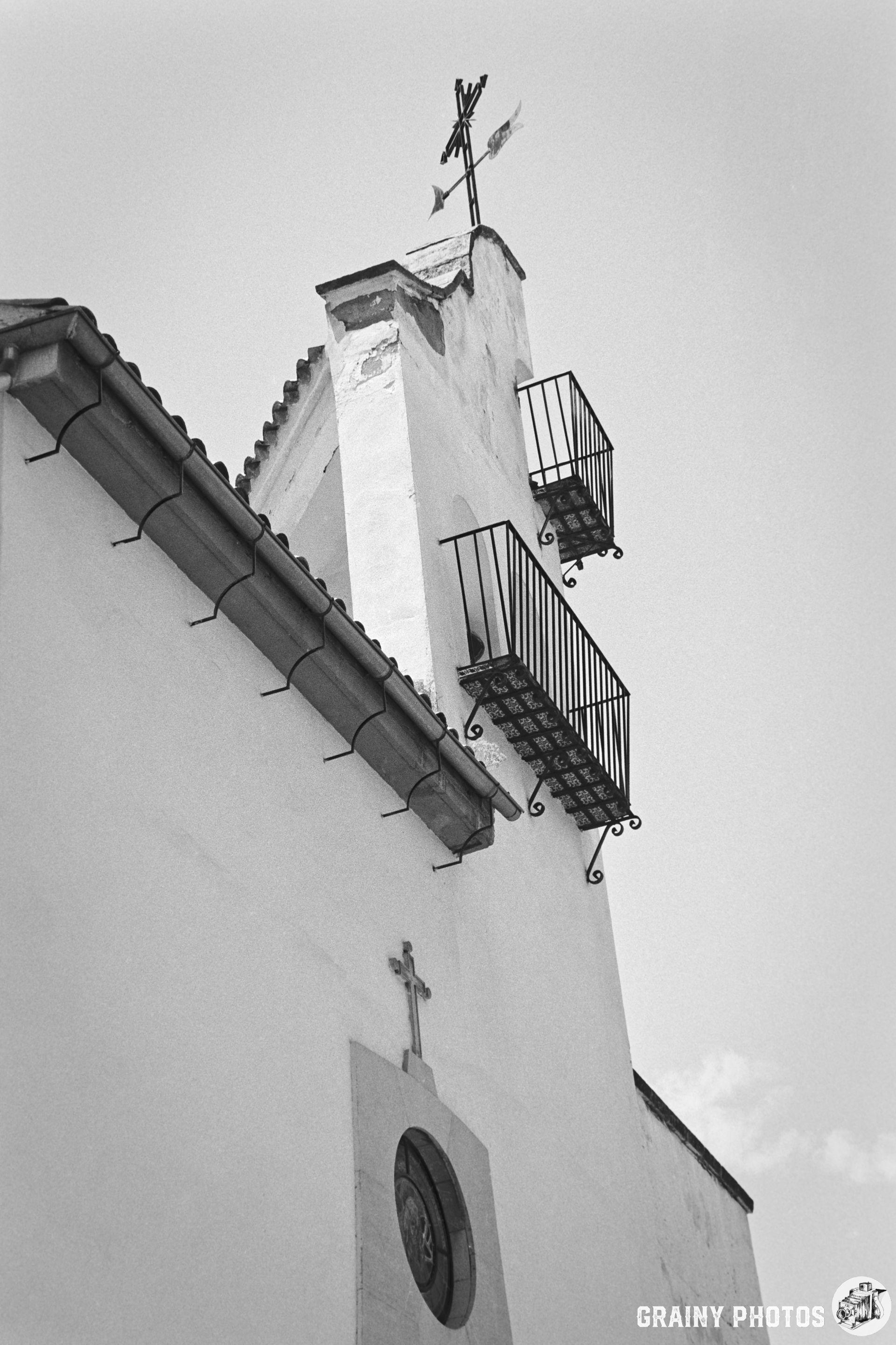 A black and white image of a church belfry featuring a cross atop the steeple and decorative balconies, highlighting its architectural details against a clear sky.