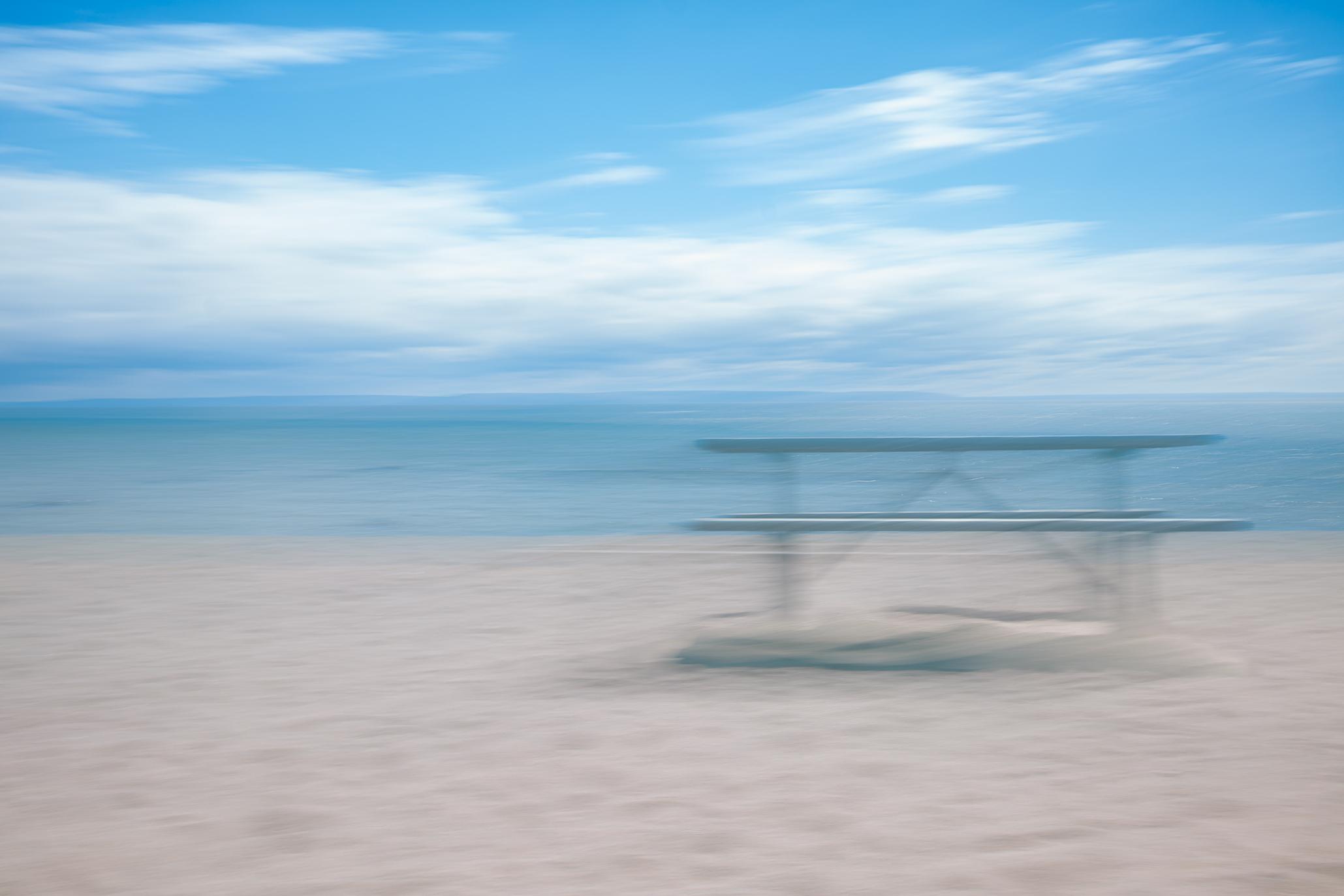 An intentional camera movement blurred photo of a picnic table on a sandy beach. In the background, a blue stretch of water and a bright blue sky with white clouds.