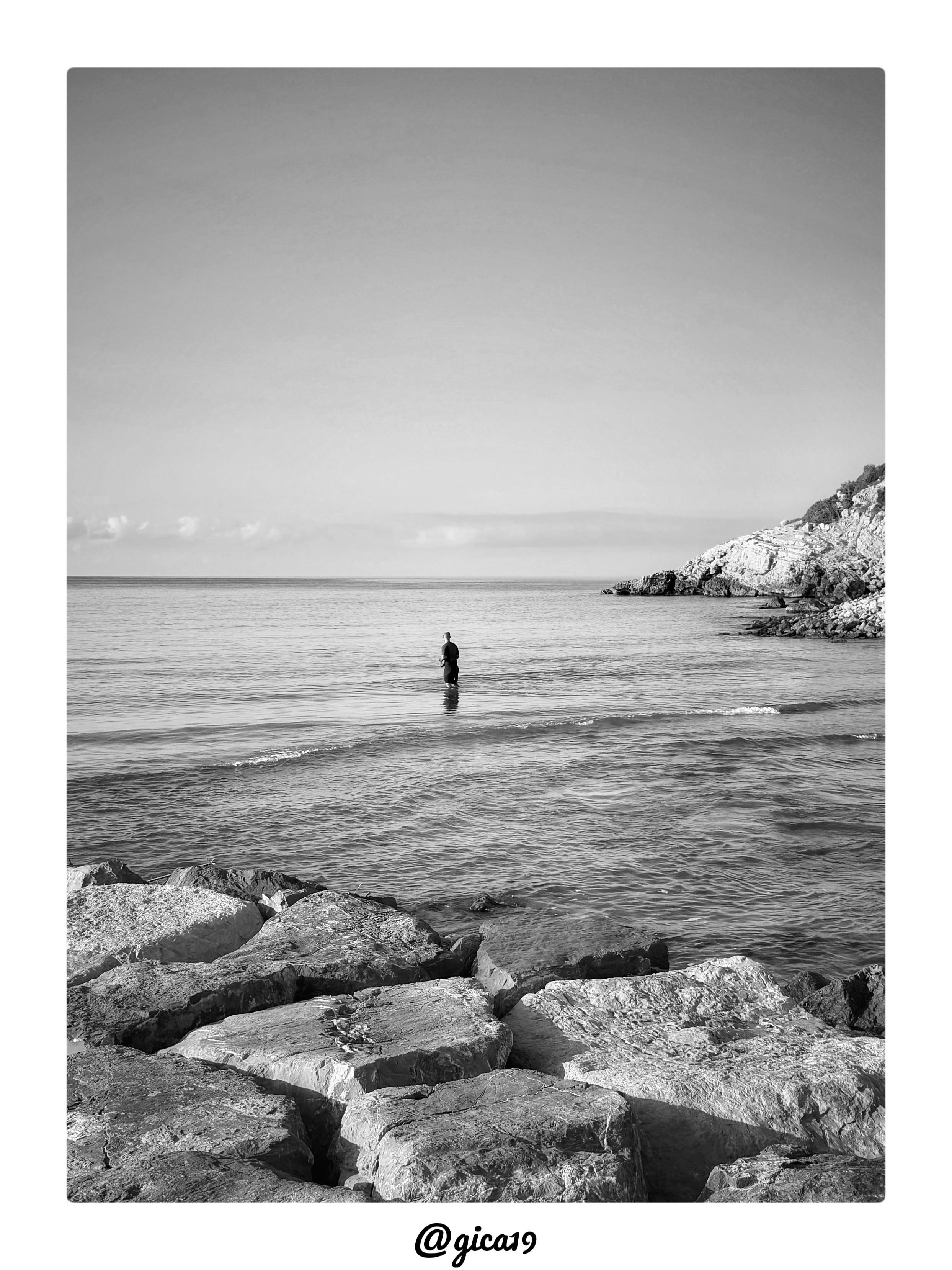 La imagen muestra una escena marina en blanco y negro tomada en la Playa de las Viudas, junto a la Playa Sur de Peñíscola, con una composición serena y minimalista, típica de la fotografía de paisaje costero.

El encuadre se divide en tres planos claramente diferenciados. En primer término, ocupando la parte inferior del cuadro, aparecen grandes bloques de roca de superficie irregular y agrietada, con vetas y texturas que el blanco y negro acentúa notablemente, resaltando el contraste entre las zonas iluminadas y las sombras entre las grietas. Este primer plano rocoso actúa como marco natural que dirige la mirada hacia el mar.

En el plano medio se extiende una superficie de agua en calma, con pequeñas olas suaves que rompen cerca de la orilla dejando una fina línea de espuma. Hacia el centro de la composición, un hombre aparece de pie dentro del agua, sumergido hasta aproximadamente la cintura, de espaldas al espectador y mirando hacia el horizonte. Su figura, oscura y silueteada cont