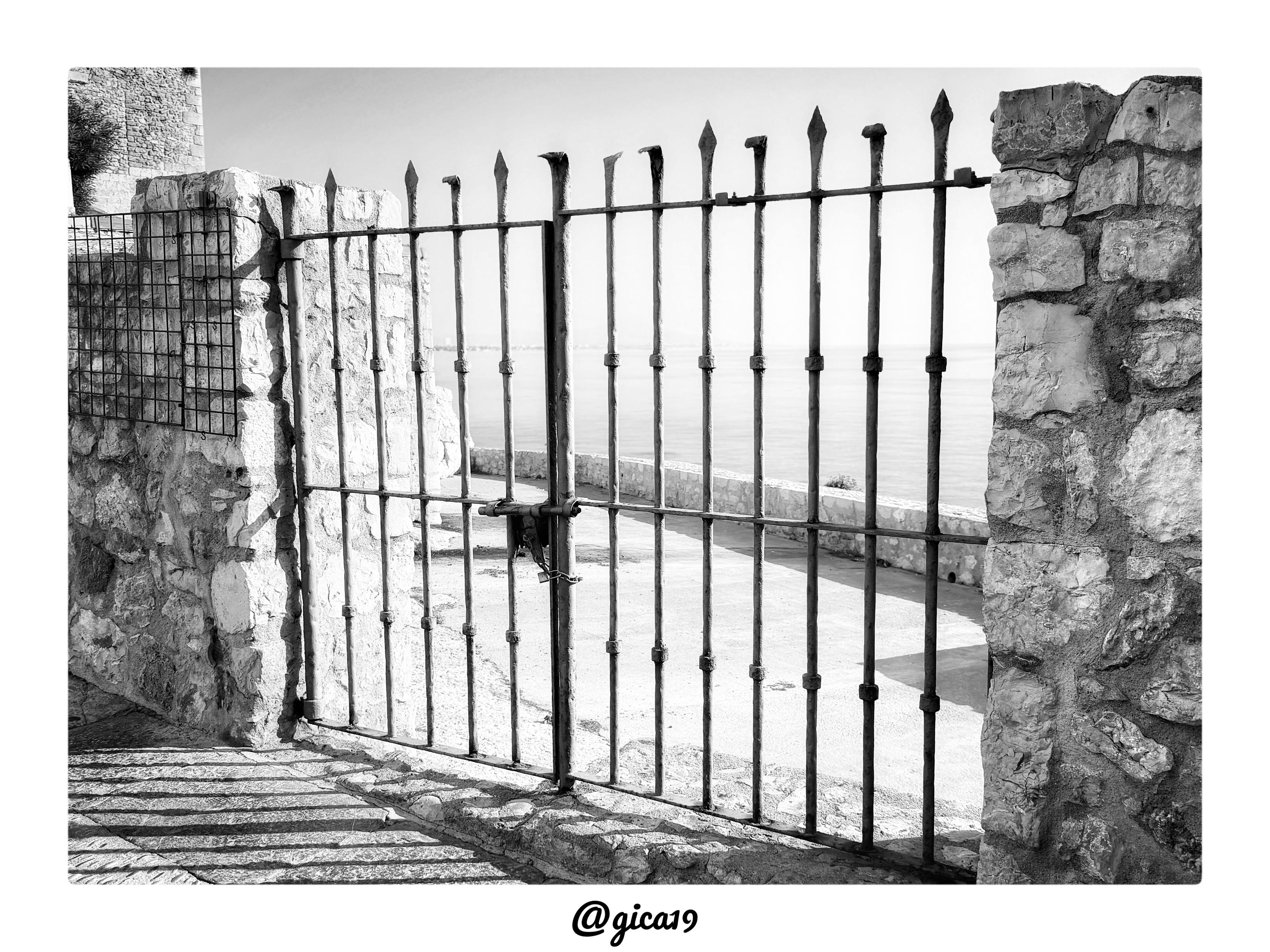 Esta es una imagen en blanco y negro que muestra una puerta de rejas de hierro en la zona antigua de Peñíscola, junto al Castillo del Papa Luna.

La fotografía está tomada desde un callejón empedrado en cuesta, encuadrando una vieja cancela metálica que separa el paso interior del pueblo de un mirador o paseo exterior junto al mar. La puerta, de hierro forjado y visiblemente oxidada, está formada por barrotes verticales rematados en punta de lanza, algunos ligeramente torcidos por el paso del tiempo, y sujeta mediante travesaños horizontales remachados. Está cerrada con un candado colocado en su punto central.

A ambos lados de la cancela se levantan dos gruesos muros de mampostería de piedra irregular, característicos de las fortificaciones y construcciones históricas del casco antiguo peñiscolano que rodea el castillo. En la esquina superior izquierda se distingue el volumen de una torre o edificación de piedra, probablemente parte del recinto amurallado o de las construcciones anexa