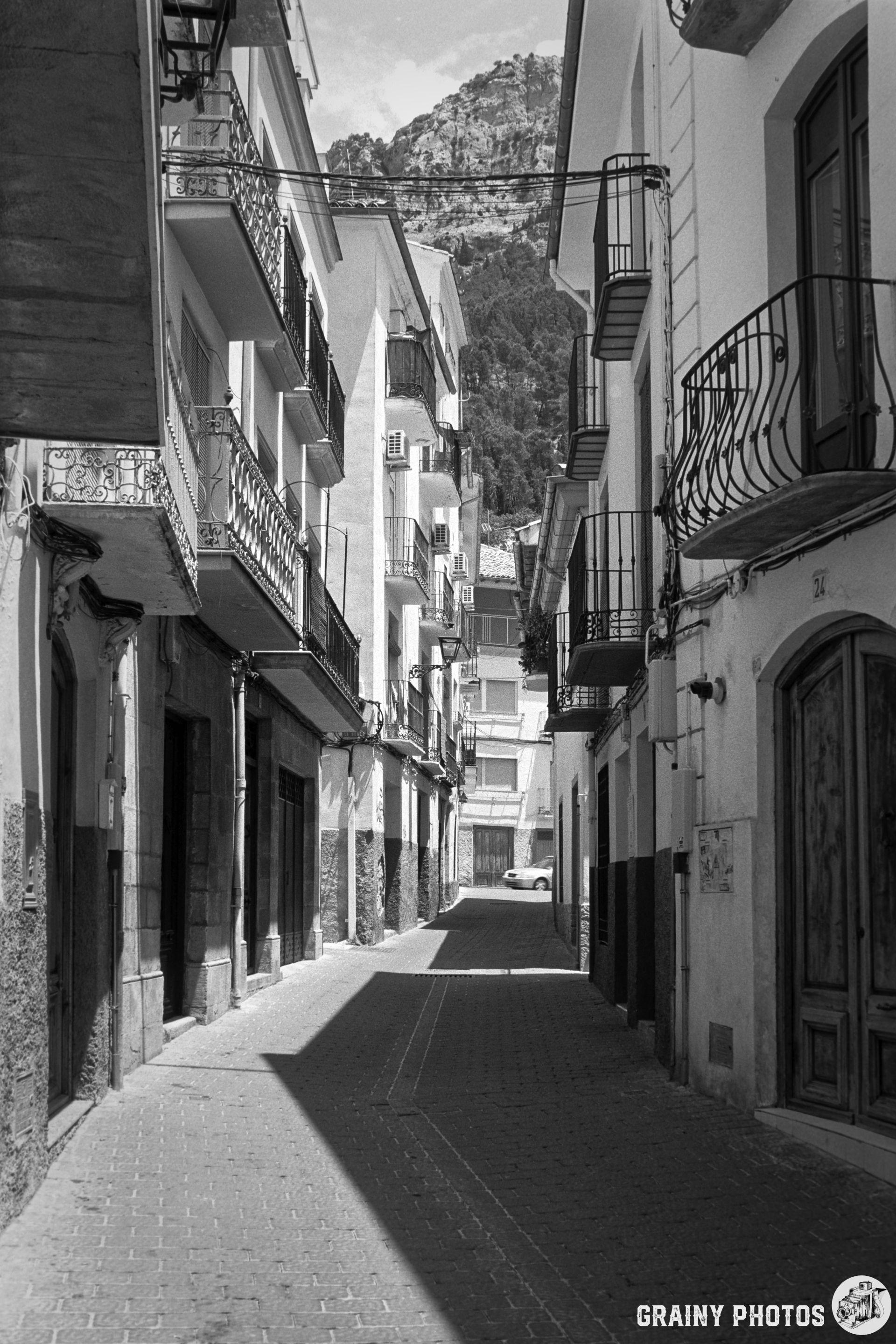 Narrow cobblestone street in Cazorla, lined with traditional buildings. Balconies overlook the alley, and a mountain is visible in the background. The scene captures a tranquil, historic atmosphere in monochrome tones.