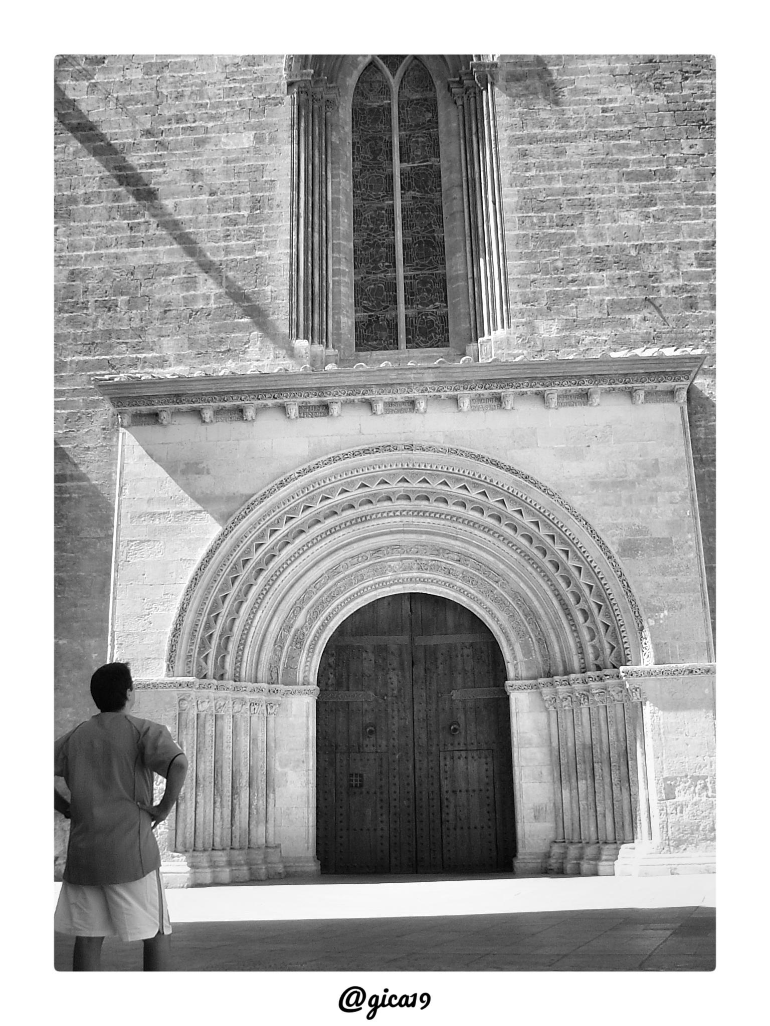 En la fotografía, en blanco y negro, se ve el lateral de la Catedral de Valencia correspondiente a su portada románica, conocida como Puerta del Palau o de la Almoina, situada junto al antiguo palacio arzobispal. Domina la composición un gran arco abocinado formado por varias arquivoltas concéntricas decoradas con motivos geométricos —zigzags, dentículos y arquillos ciegos—, apoyadas sobre columnas con capiteles esculpidos, un esquema típico del románico de influencia lemosina que se levantó pocos años después de la conquista cristiana de la ciudad en 1238. Sobre el arco se recorta una gran ventana ojival geminada con vidriera, y todo el conjunto está construido en sillería de piedra caliza, con la superficie desgastada por el paso del tiempo. Las puertas de madera, robustas y con clavazón metálica visible, permanecen cerradas en el momento de la toma. En primer plano, de espaldas y a la izquierda del encuadre, aparece una persona con camiseta y pantalón corto observando el edificio, l