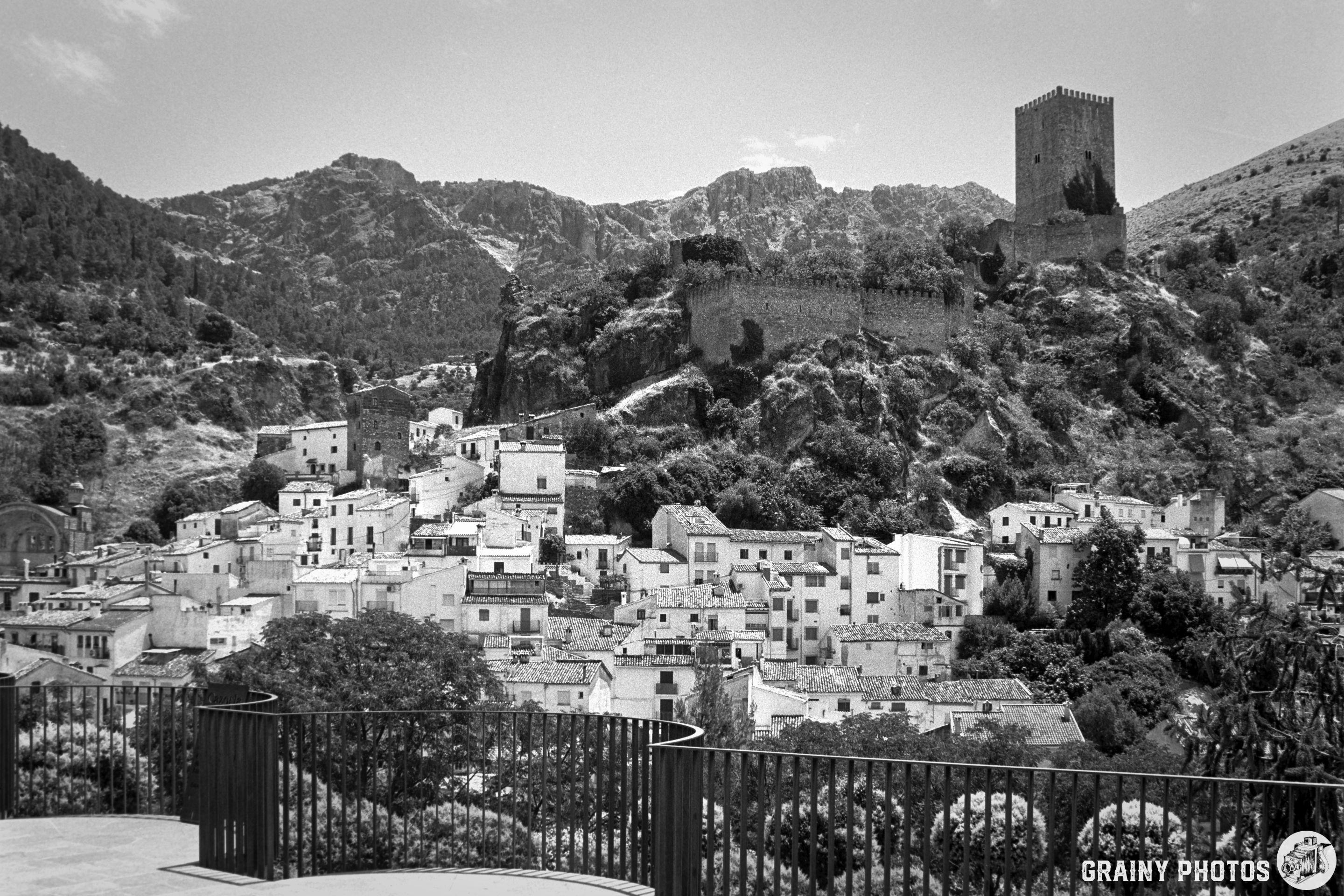 A scenic black-and-white view the old part of Cazorla surrounded by mountains, featuring traditional whitewashed houses and a prominent castle tower overlooking the landscape.