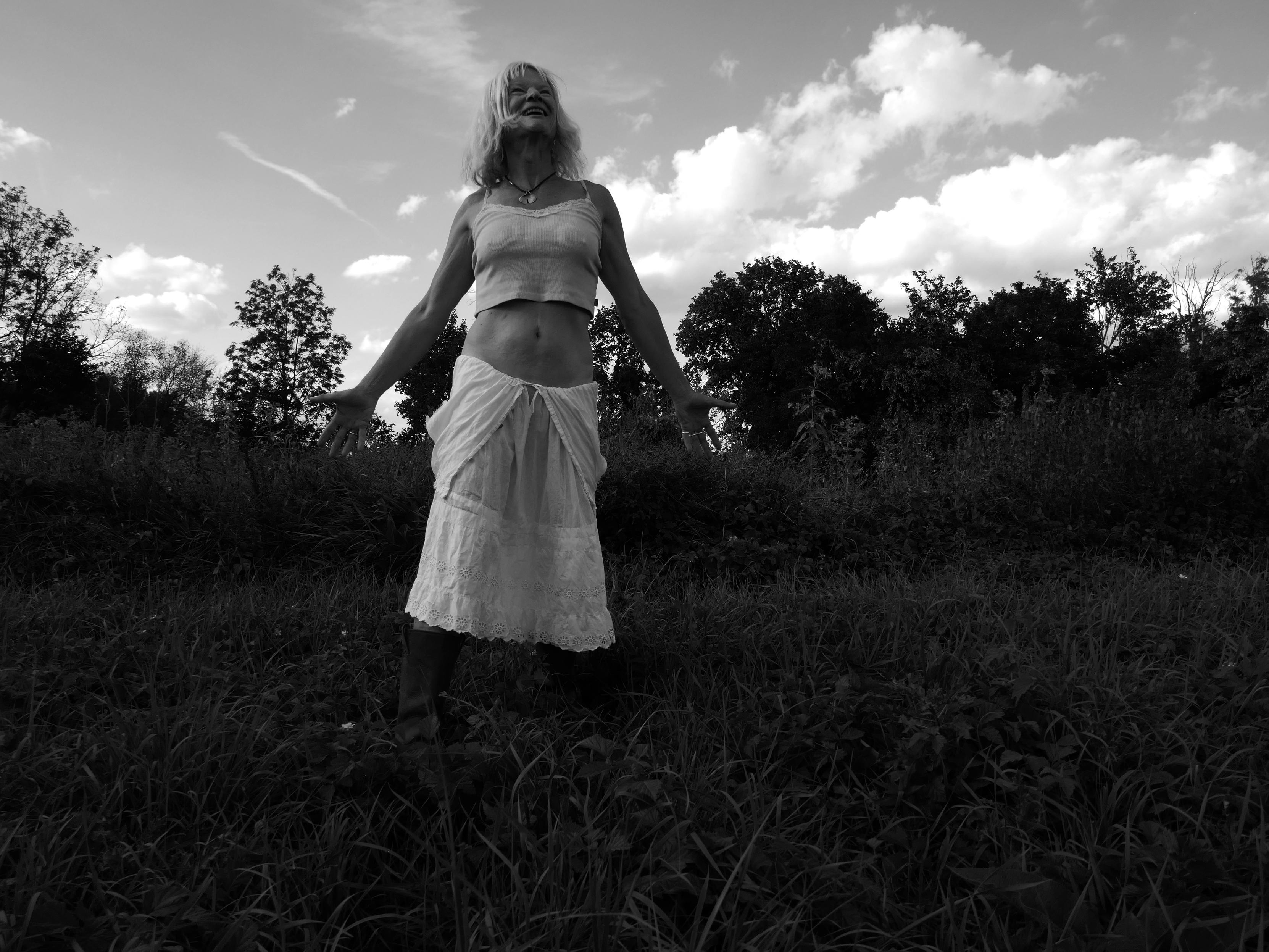 a blond woman wearing a white skirt riding low on her hips, and a slightly darker cropped top. She looks into tha sky, spreads her arms low, like graating the clouds and looks into ths sky. She stands off center in a dark meadow, with dark young trees surrounding her. There are some dramatic white clouds in a grex sky. The picture is black and white.
