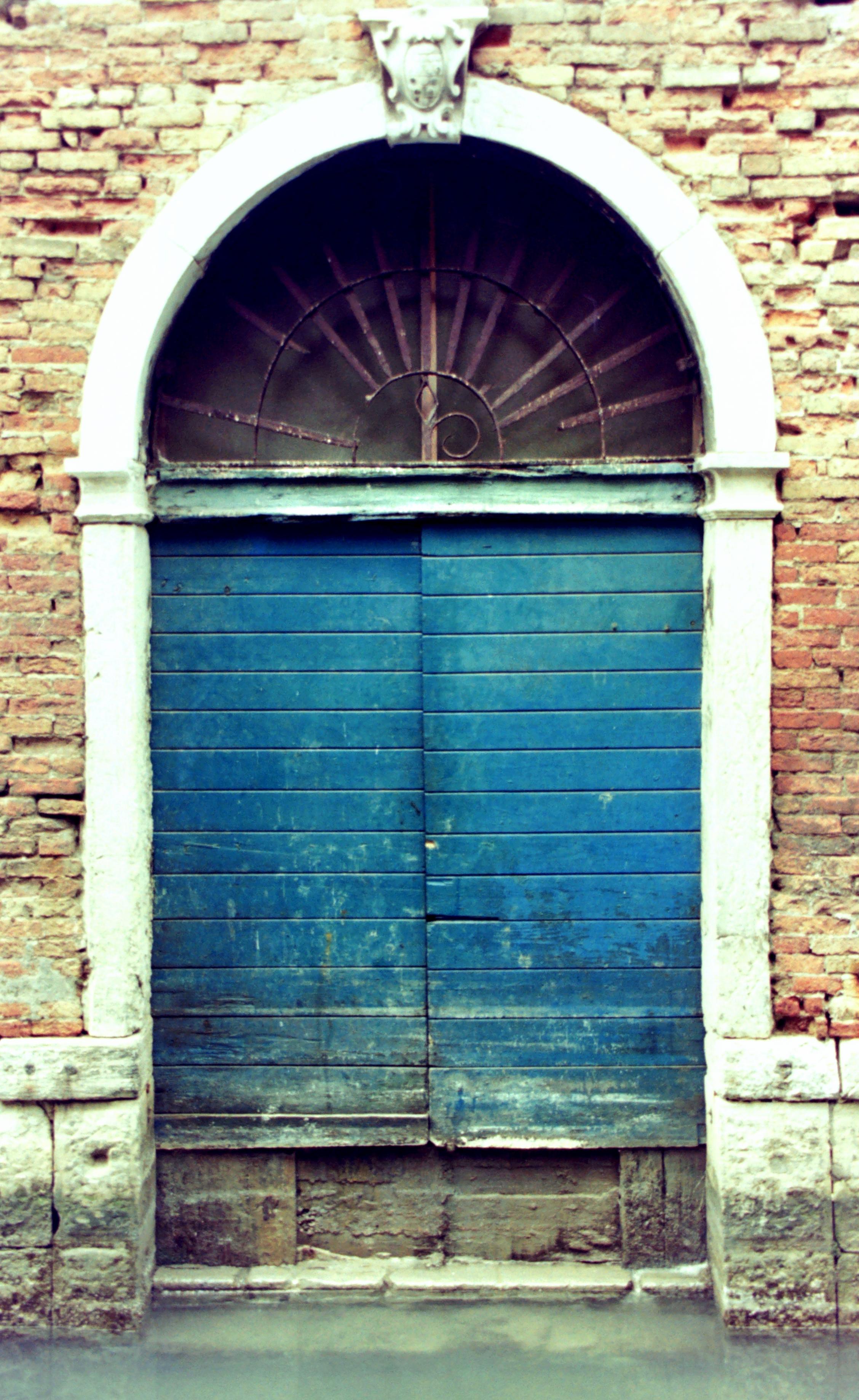 Analog color photograph in 3:4 portrait format, from the front view of the entrance to a house on a canal in Venice, in an old brick building with stone blocks at its base, eroded and darkened by water. The doorway, constructed with horizontal wooden panels painted dark blue, is flanked by two white stone pillars with capitals supporting a semicircular arch of the same stone. Below the arch is a worn, radially shaped, semicircular iron grille. A small stone shield crowns the arch.
