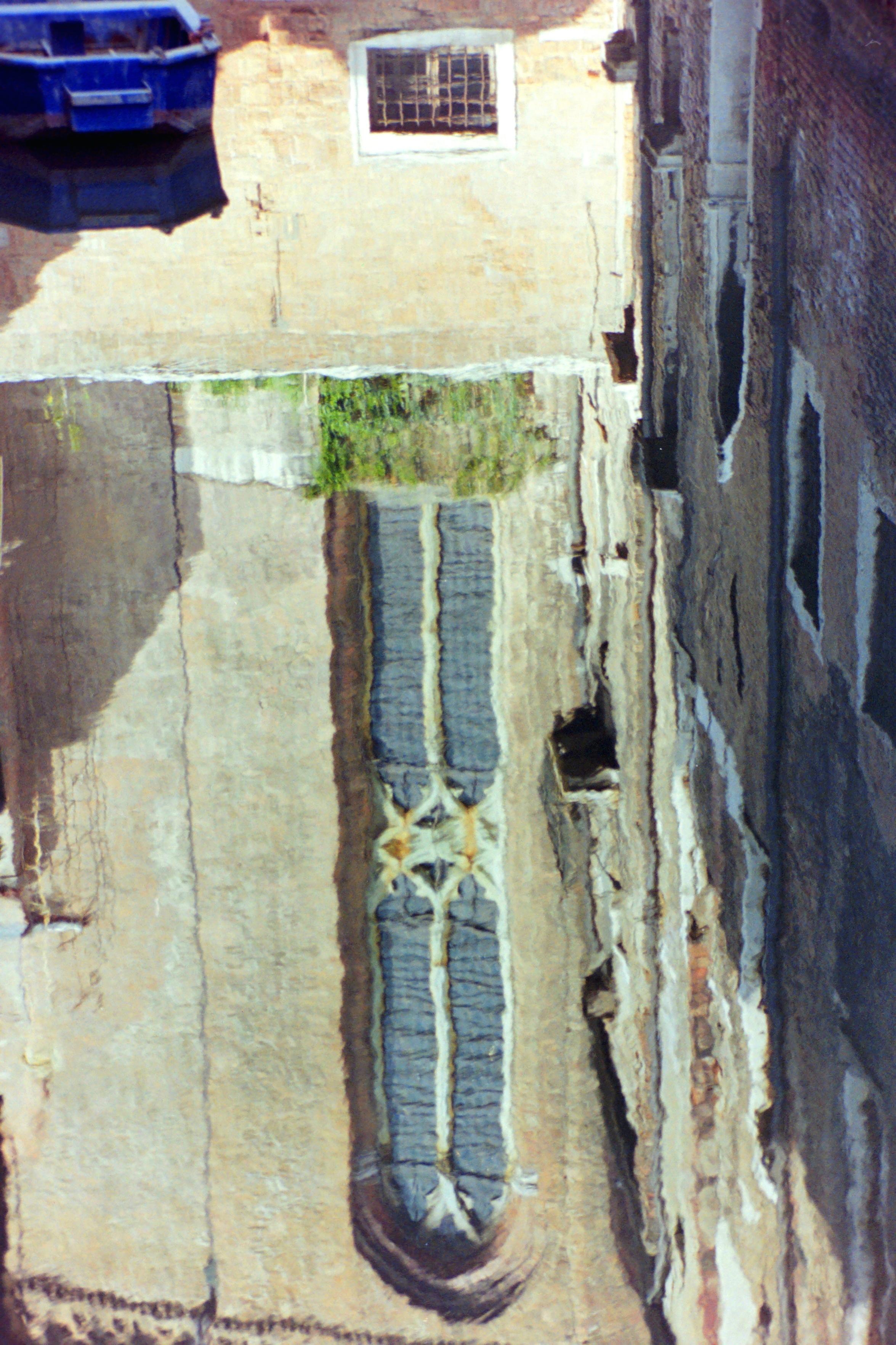Analog color photograph in 2:3 portrait format of the reflection, in the calm waters of a Venetian canal, of a facade with a large Venetian Gothic-style window. Also reflected are a small rectangular, barred window and the facade of the adjacent building, covered in windows. 
Part of a small boat appears in the upper left.