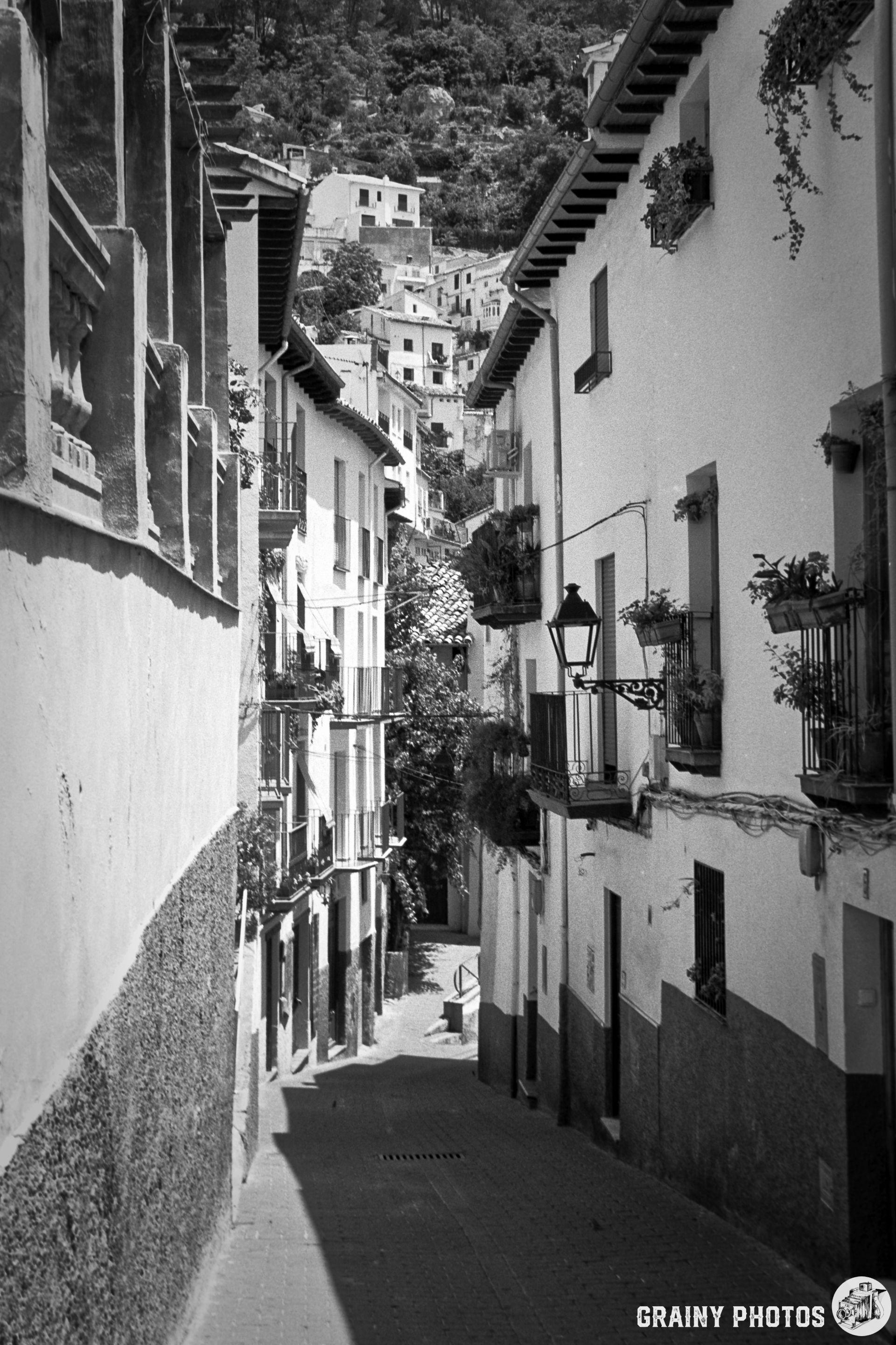 A narrow, winding street lined with whitewashed houses featuring balconies adorned with plants. The scene is captured in black and white, with a hillside visible in the background, showcasing traditional architecture and a tranquil atmosphere.A narrow, winding street lined with whitewashed houses featuring balconies adorned with plants. The scene is captured in black and white, with a hillside visible in the background, showcasing traditional architecture and a tranquil atmosphere.