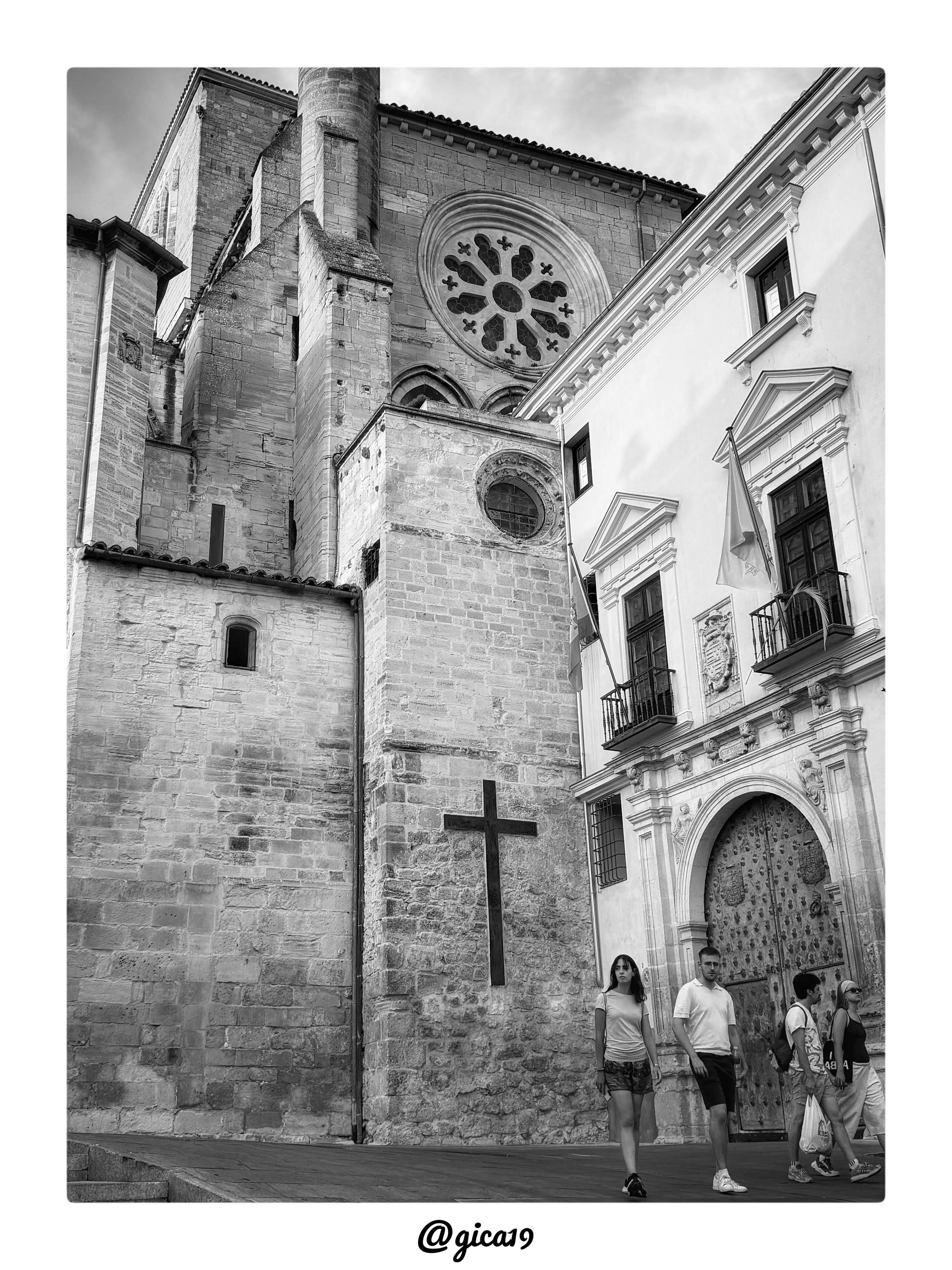 La fotografía en blanco y negro capta el lateral de la Catedral de Santa María y San Julián de Cuenca, con su fachada de sillería de piedra caliza cortada en grandes bloques irregulares que revelan el paso del tiempo sobre la construcción gótica.

En la parte superior destaca el gran rosetón, elemento que domina visualmente toda la composición. Presenta un diseño radial con pétalos lobulados dispuestos en torno a un núcleo circular central, encerrado en un marco de piedra labrada de forma ligeramente ovalada por el ángulo de la toma. Su tracería calada, típica del gótico, contrasta por la profundidad de sus huecos oscuros frente a la superficie más clara de la piedra que lo rodea, y corona de forma casi ceremonial el resto de la fachada. Justo debajo se adivinan los arranques de un triforio o galería de arcos ciegos, y algo más abajo un segundo óculo circular de menor tamaño, más sencillo en su decoración, que repite el motivo circular a escala reducida.

A la izquierda se eleva una to