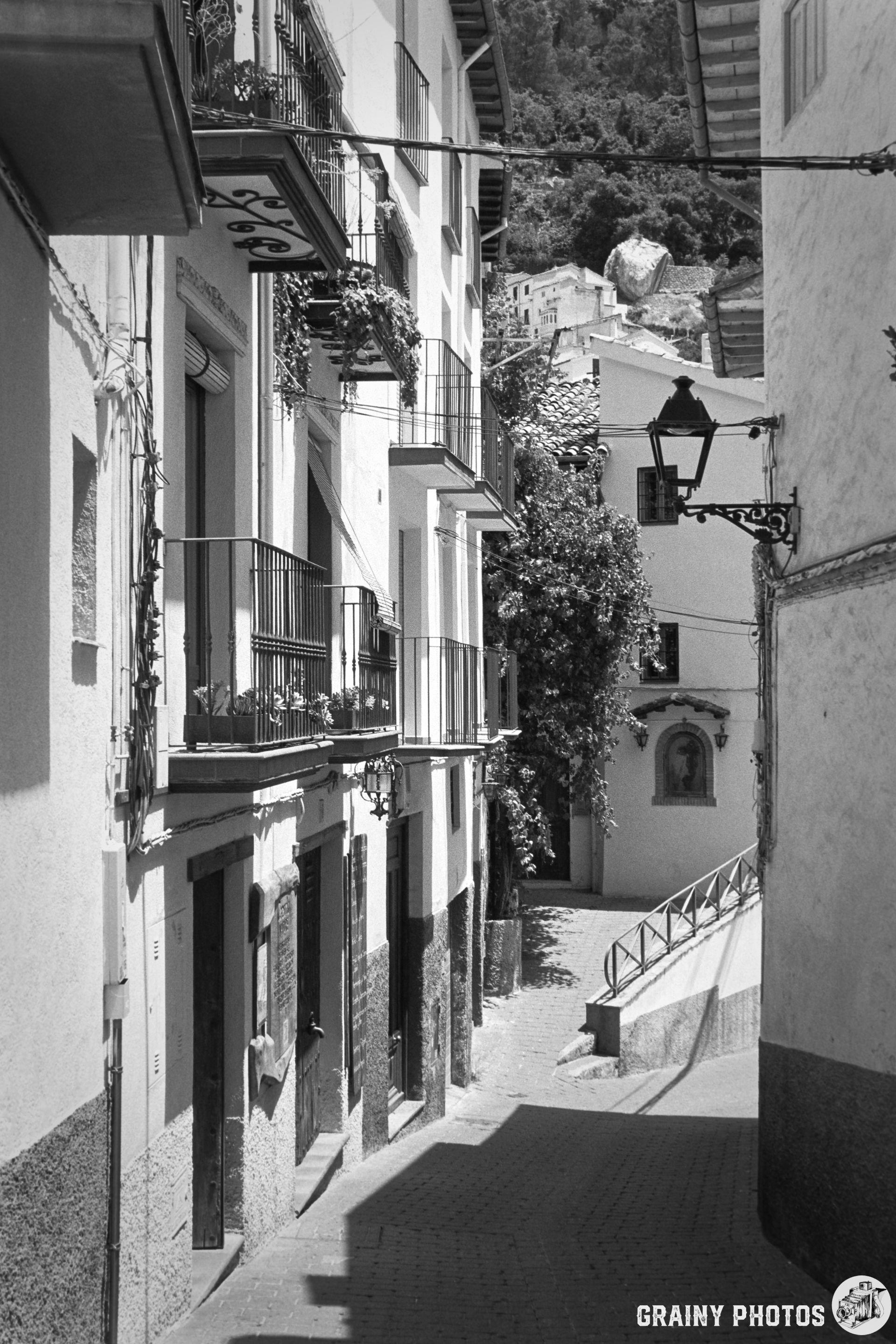 A narrow, cobblestone street lined with whitewashed buildings featuring balconies adorned with plants. The scene is set in a charming village, highlighted by a lantern and a distant rocky hill under a clear sky. The image is in black and white.