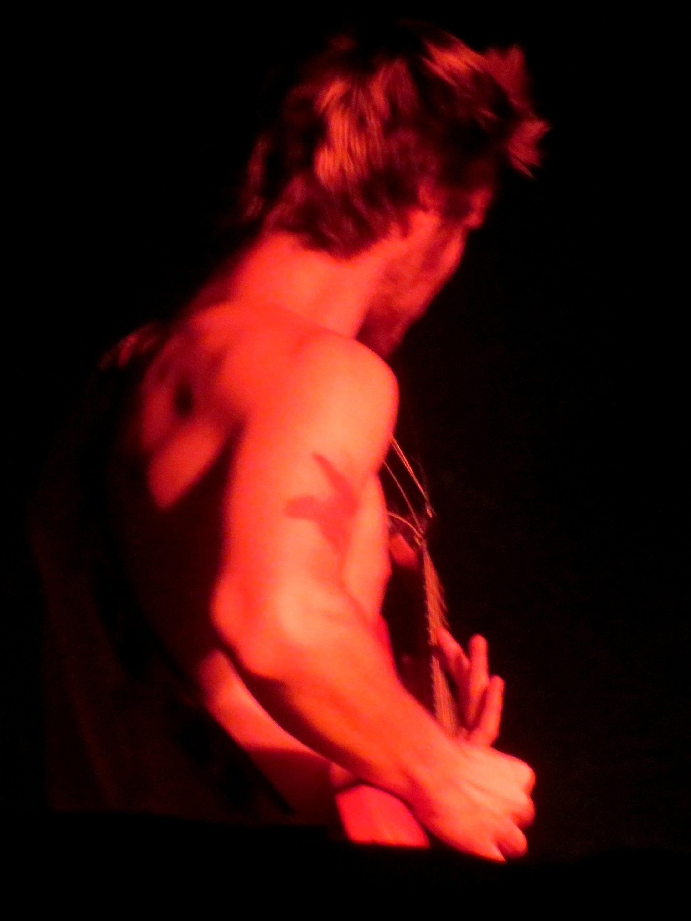 Photograph of a member of a musical group playing electric guitar at a concert with his torso bare and his arm tattooed under a reddish light.